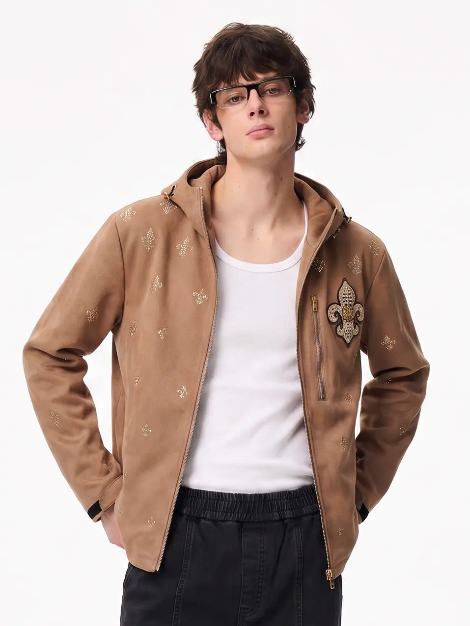A man wearing a tan zip-up hooded jacket with gold fleur-de-lis patterns and a gold crest patch on the chest, layered over a white tank top and black pants, against a plain white background. TCH Rhinestone Fleur-de-Lis Hooded Jacket