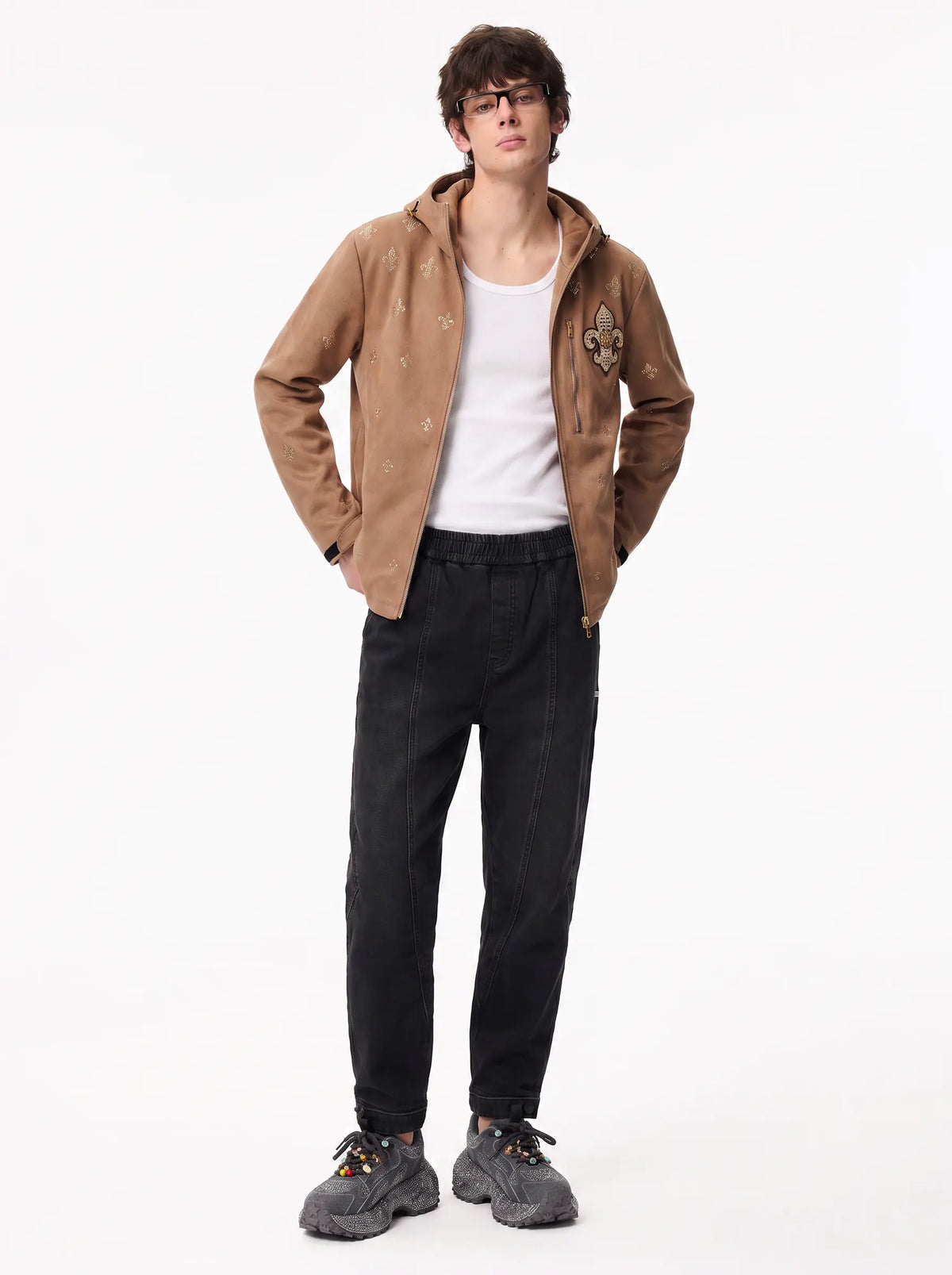 A male model wearing a brown hooded jacket with embroidered details, a white tank top, black pants, and gray sneakers, standing against a plain white studio background. TCH Rhinestone Fleur-de-Lis Hooded Jacket