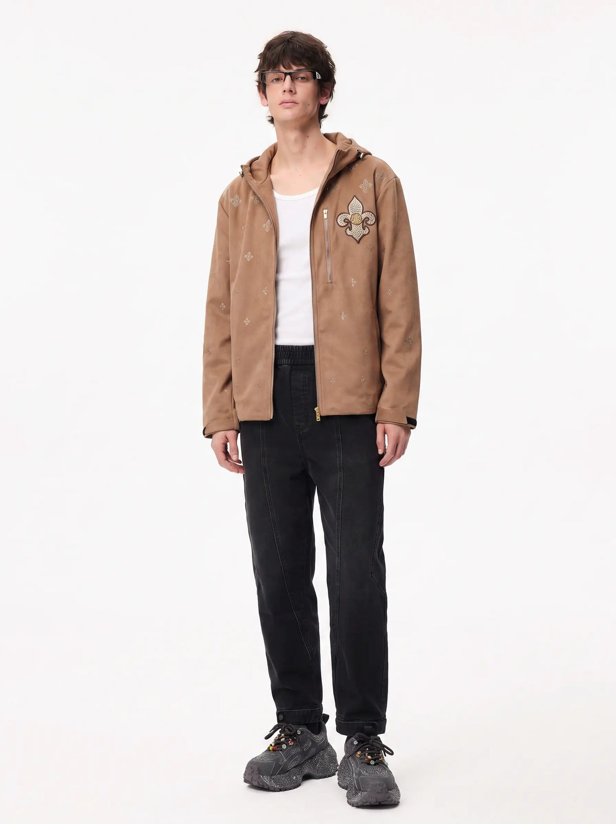 A man wearing a tan hooded jacket with a fleur-de-lis patch on the chest, styled over a white t-shirt, black jeans, and gray sneakers, standing against a plain white studio background. TCH Rhinestone Fleur-de-Lis Hooded Jacket
