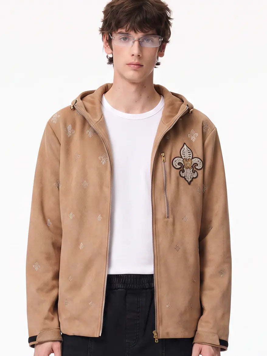 A male model wearing a tan zip-up hooded jacket with embroidered fleur-de-lis motifs, layered over a white t-shirt and black pants, against a plain white background. TCH Rhinestone Fleur-de-Lis Hooded Jacket