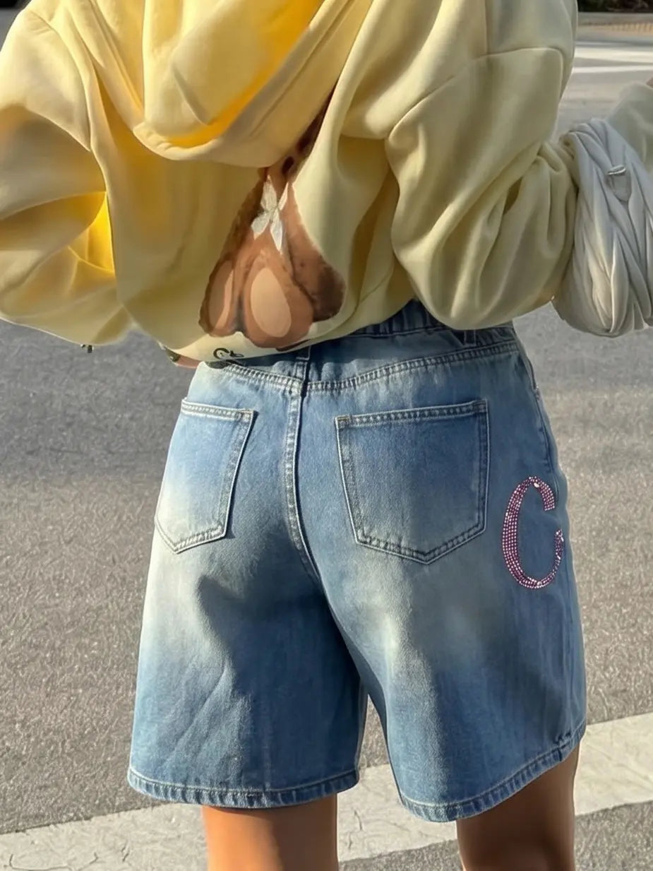 Person wearing a yellow hoodie and blue denim shorts with a logo on a street. TCH Rhinestone Floral Logo Wide-Leg Denim Shorts