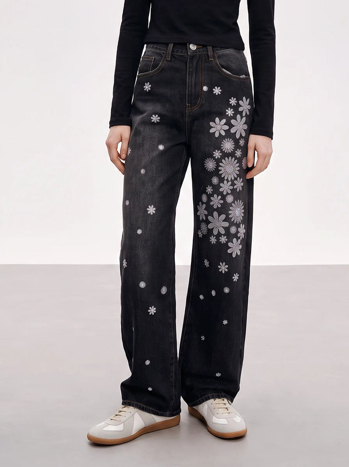 A model is shown from the waist down wearing a pair of wide-leg black denim jeans decorated with white embroidered flowers and dots, paired with a black long-sleeve top and white sneakers. TCH Rhinestone Floral Wide-Leg Jeans