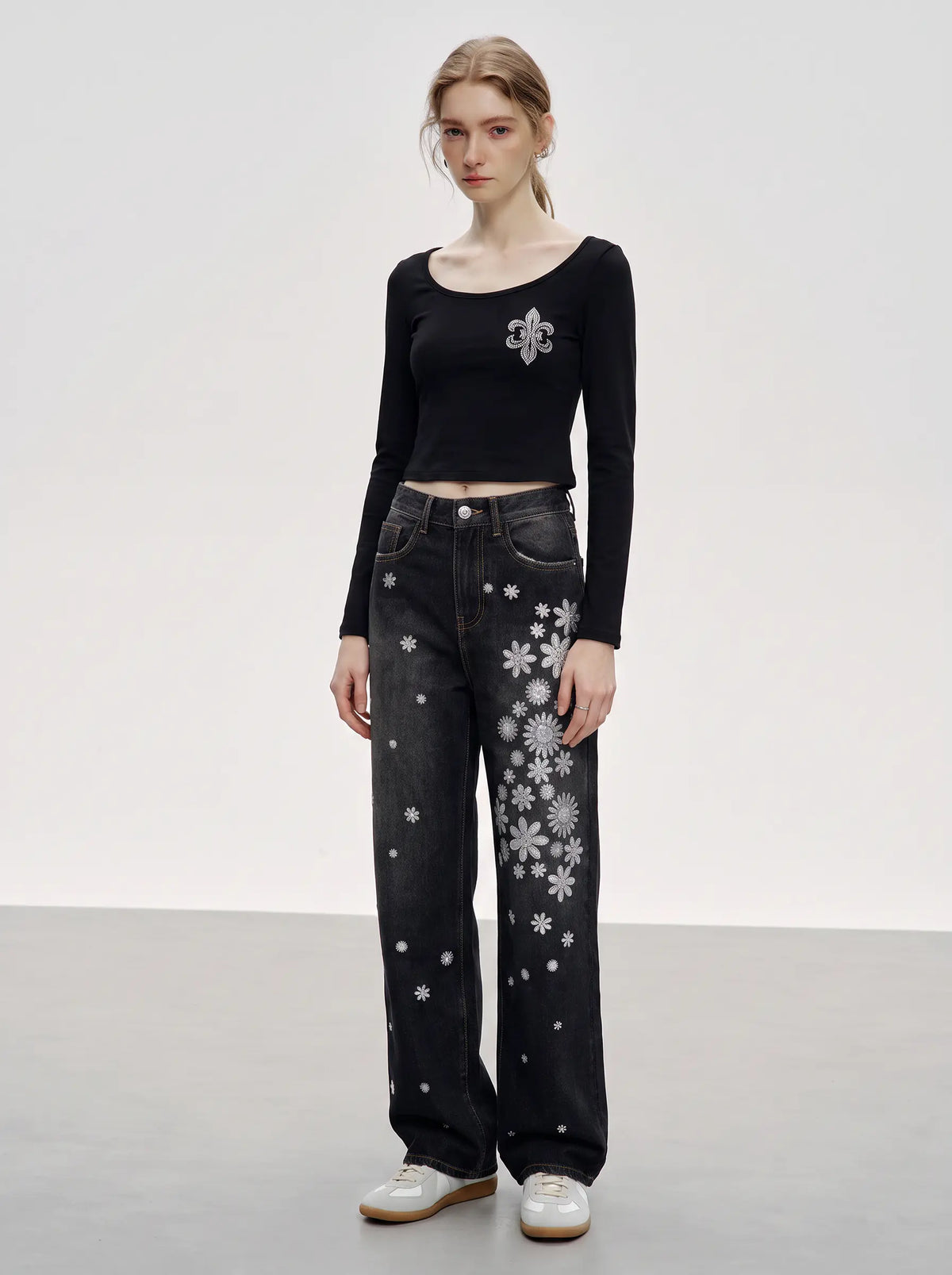 A model stands in a studio wearing a black long-sleeve crop top with a silver floral applique on the chest, paired with black wide-leg jeans featuring white snowflake patterns, and white leather shoes. TCH Rhinestone Floral Wide-Leg Jeans