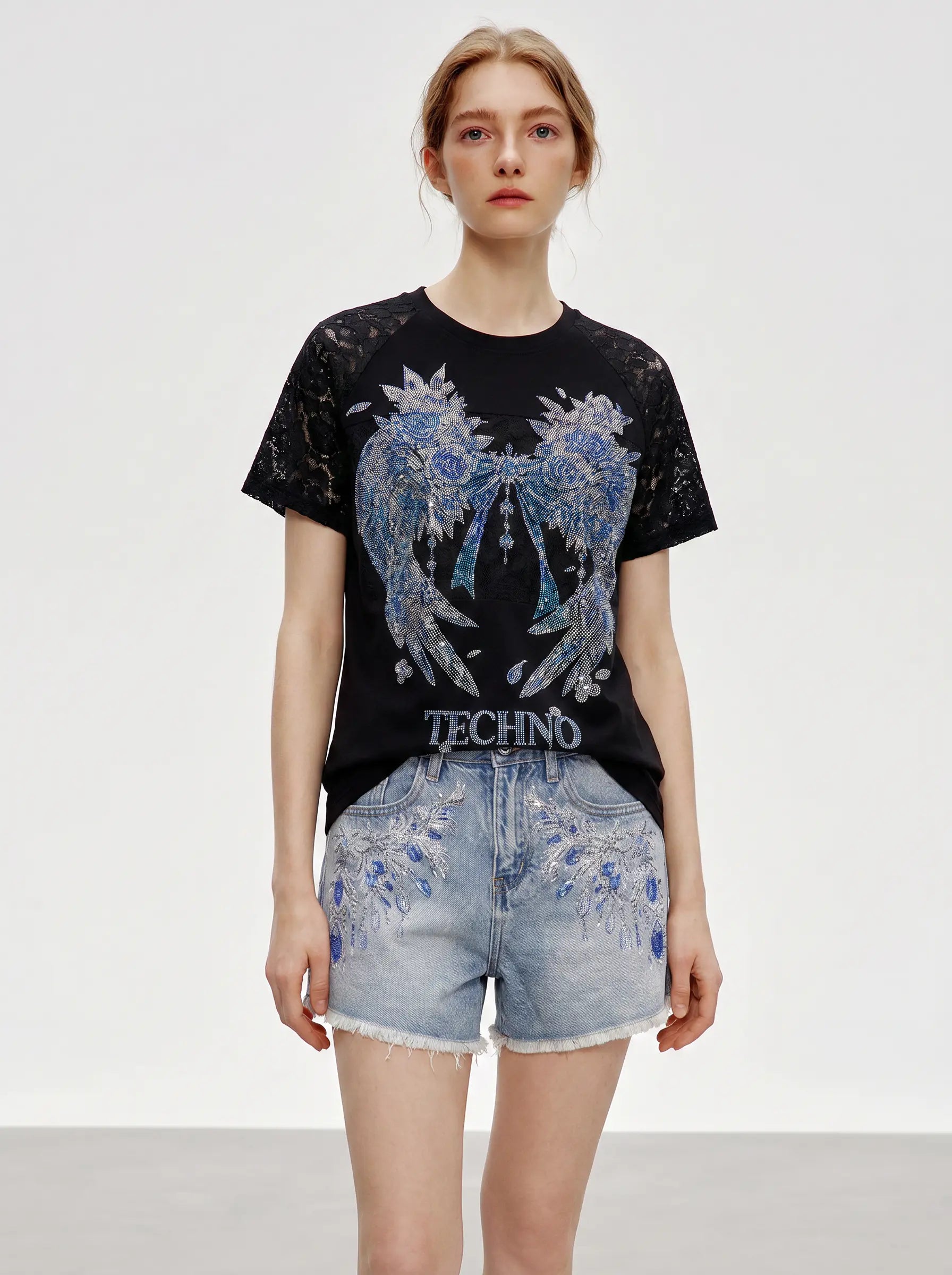 A model wearing a black short-sleeve Techno brand t-shirt with a large blue and white angel wing graphic and light-wash denim shorts with blue floral embroidery, set against a plain light gray studio background. TCH Rhinestone Floral Wing Techno Graphic T-Shirt