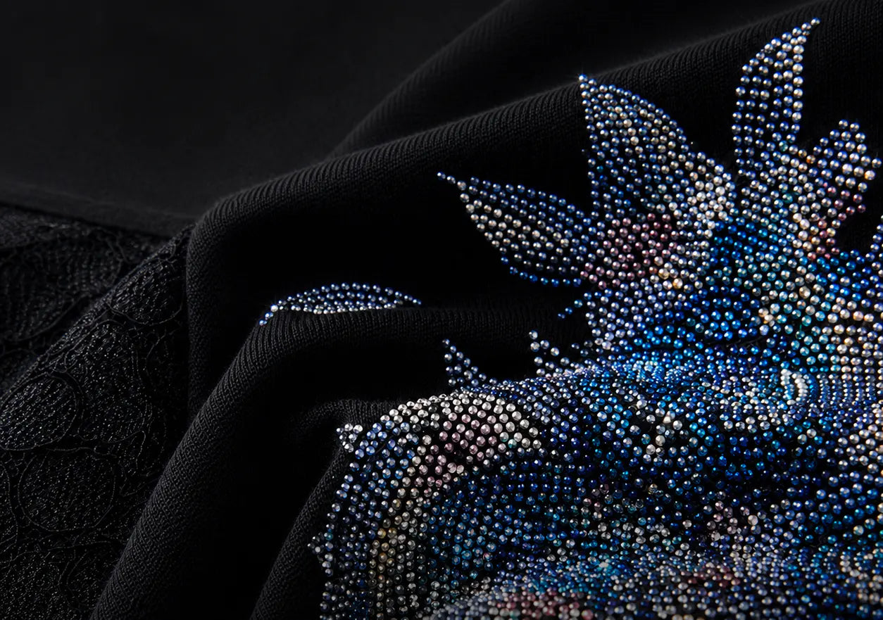 Close-up of a black fabric garment featuring an intricate, beaded floral or leaf motif in shimmering blue, silver, and iridescent tones. TCH Rhinestone Floral Wing Techno Graphic T-Shirt