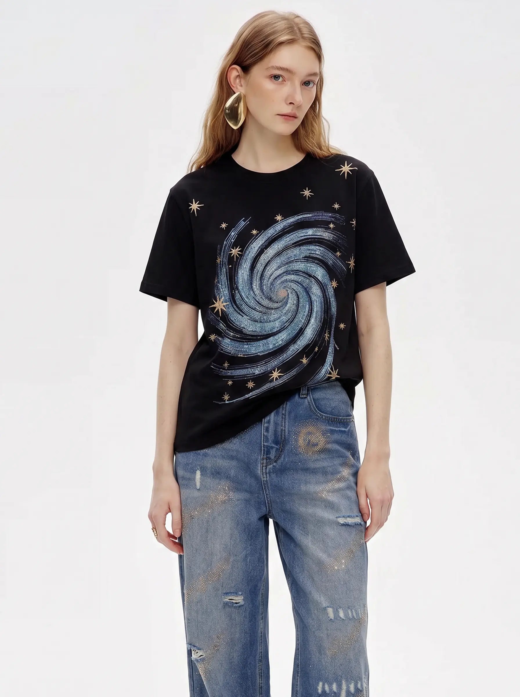A model wearing a black short-sleeve t-shirt with a blue and silver galaxy spiral graphic and gold star embellishments, paired with distressed blue jeans, against a plain white studio background. TCH Rhinestone Galaxy Spiral Graphic Black T-Shirt