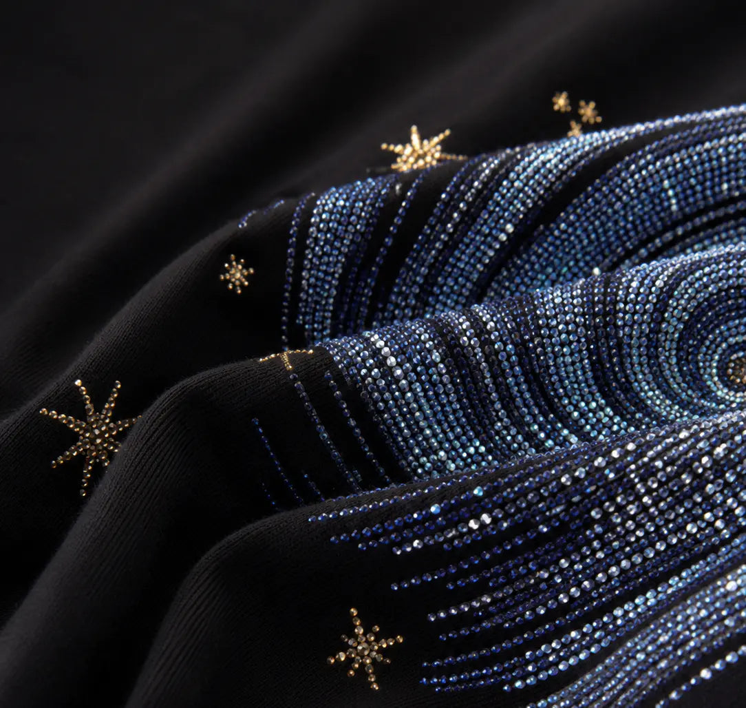 Close-up of a black fabric garment featuring intricate blue beaded swirl patterns and small gold embroidered star accents. TCH Rhinestone Galaxy Spiral Graphic Black T-Shirt