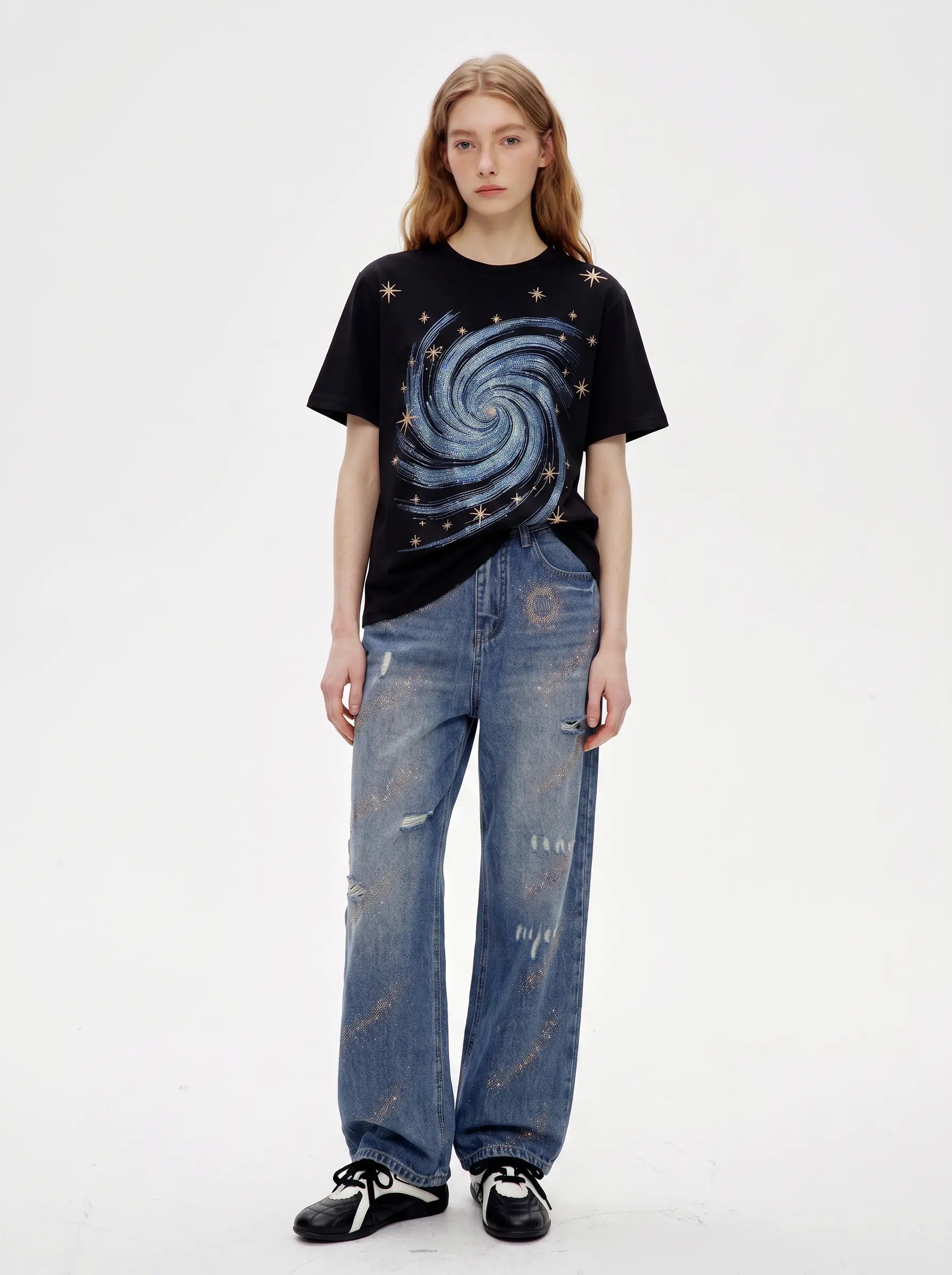 A model stands against a plain white studio background wearing a black graphic T-shirt with a blue spiral galaxy print, distressed blue jeans, and black-and-white sneakers. TCH Rhinestone Galaxy Spiral Graphic Black T-Shirt