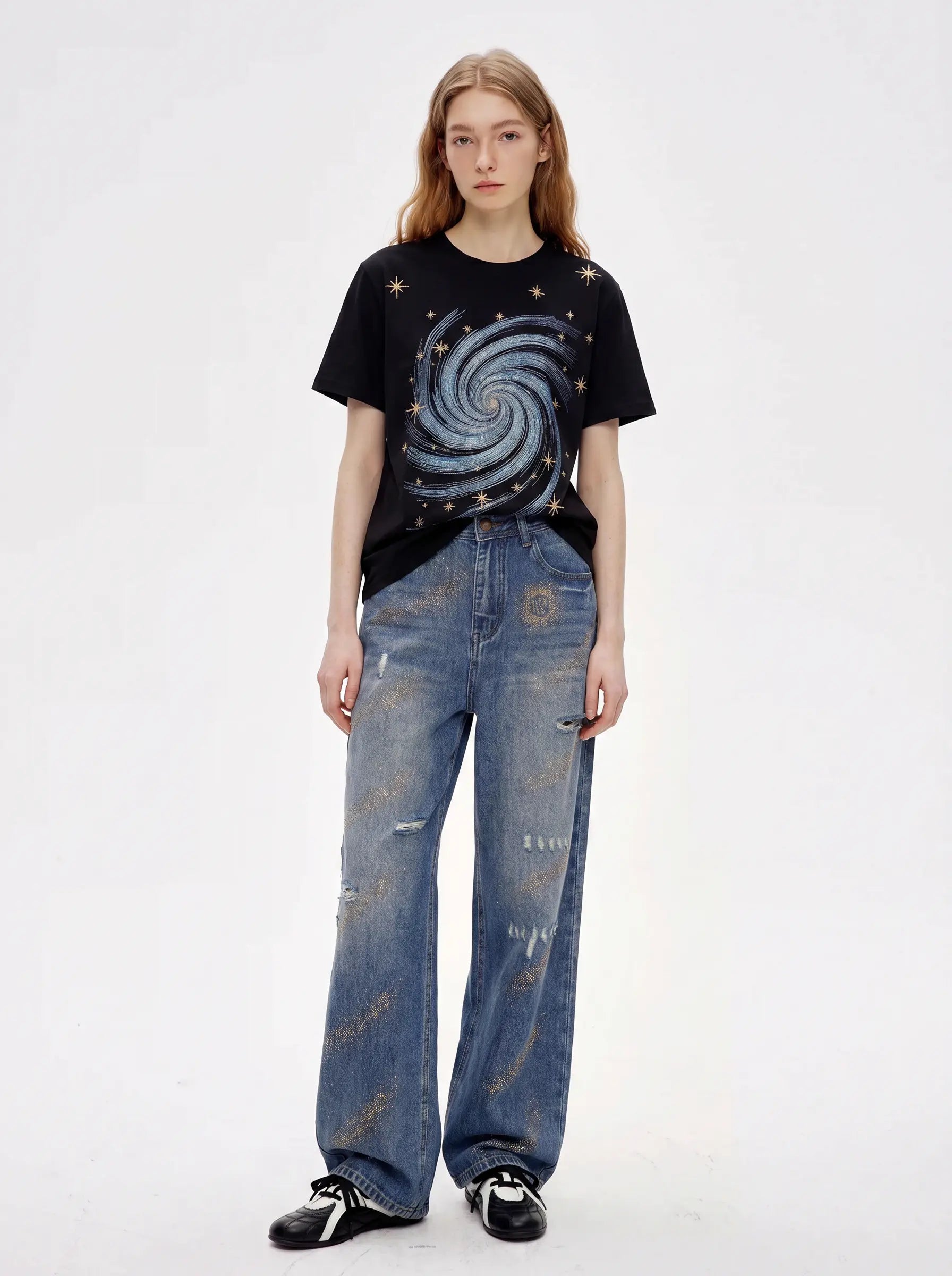 A model stands against a plain white studio background wearing a black graphic T-shirt with a blue spiral galaxy print and distressed blue jeans. TCH Rhinestone Galaxy Spiral Graphic Black T-Shirt