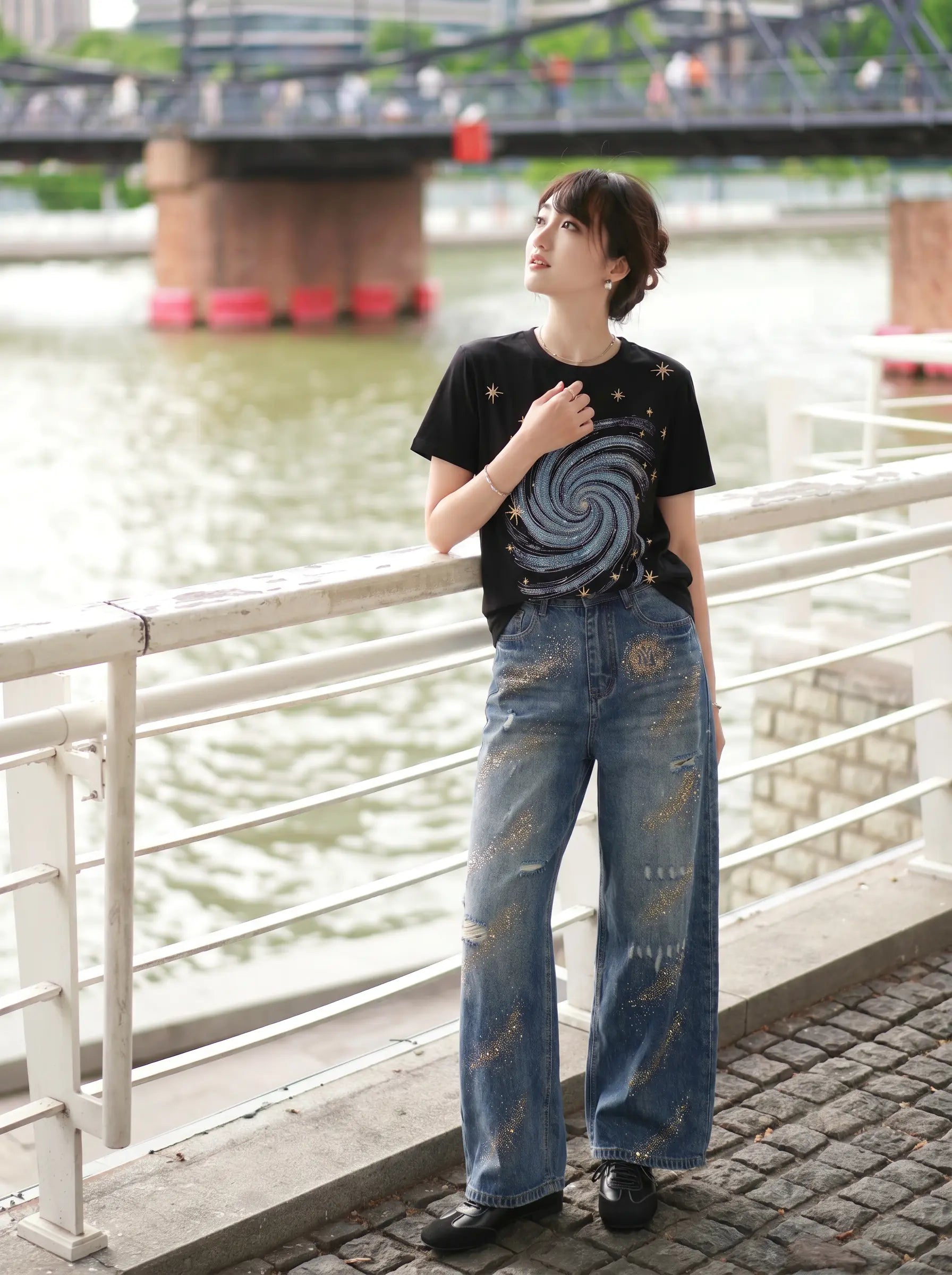 A woman stands outdoors by a river railing wearing a black graphic T-shirt with a spiral print and distressed blue wide-leg jeans. TCH Rhinestone Galaxy Spiral Graphic Black T-Shirt