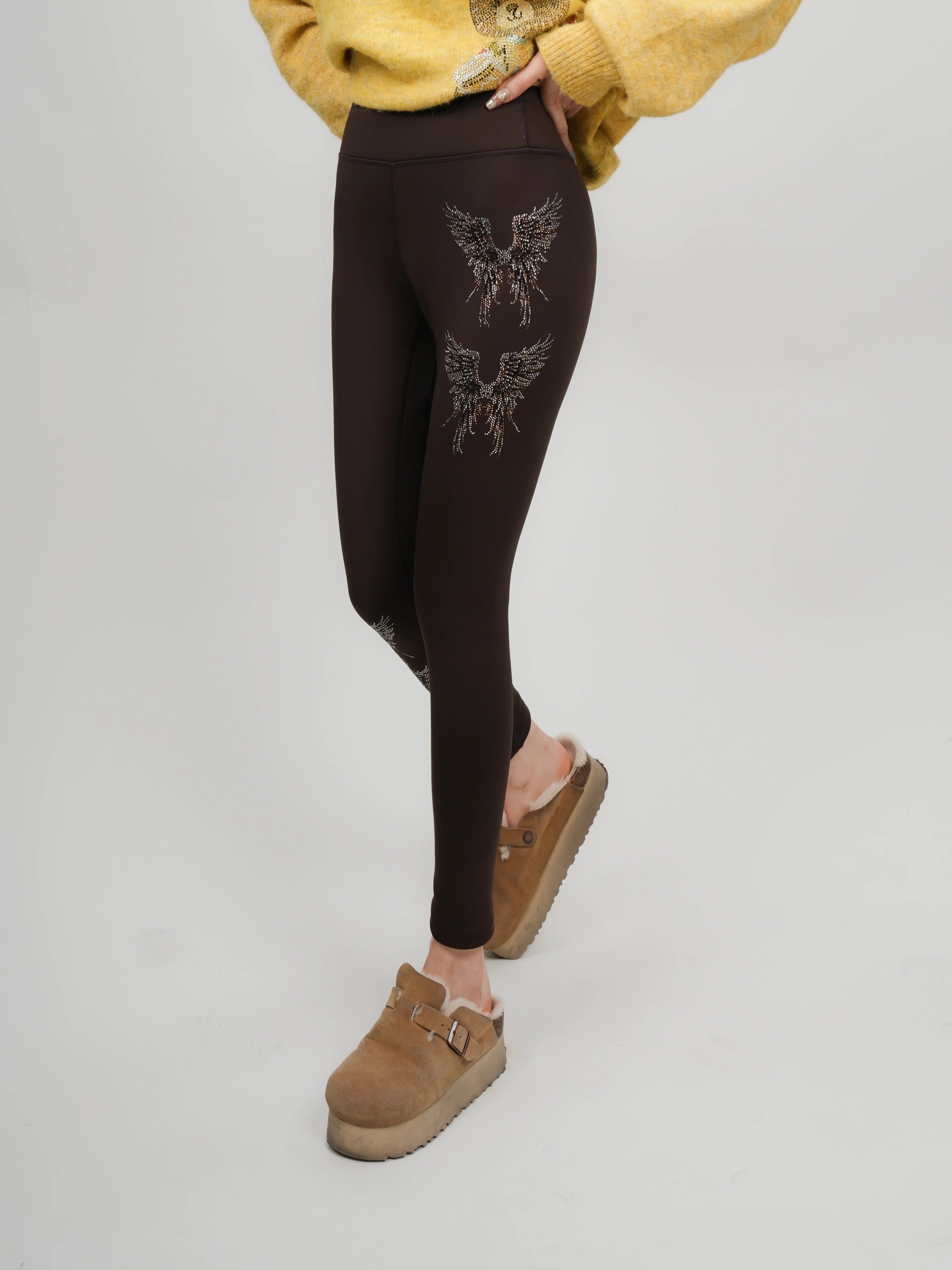 Person wearing brown leggings with a decorative design, yellow sweater, and brown shoes on a light gray background, TCH Rhinestone Golden Wings Black High-Waist Leggings