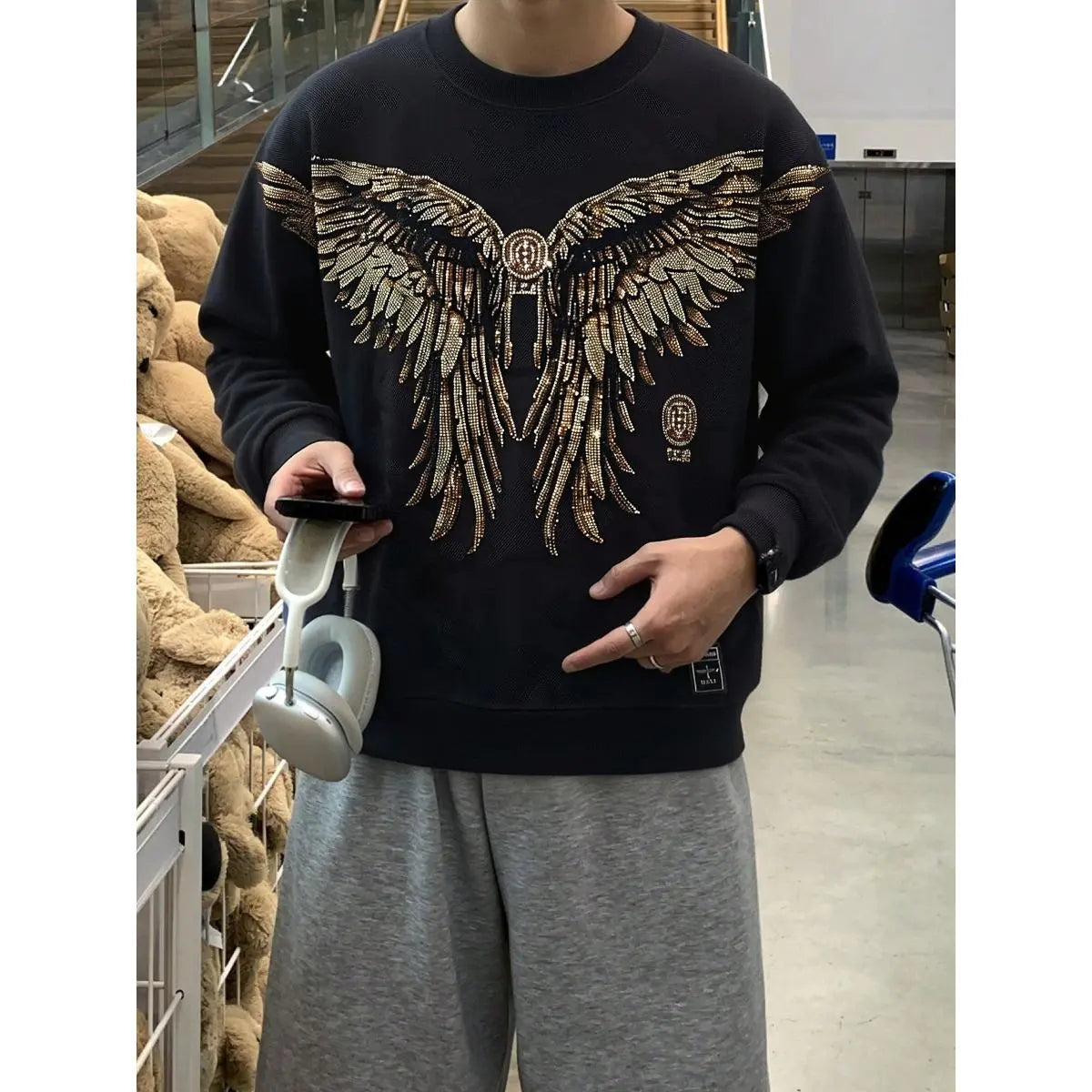 Person wearing a black sweatshirt with a large winged design in a store setting. TCH Rhinestone Golden Wings Sweatshirt