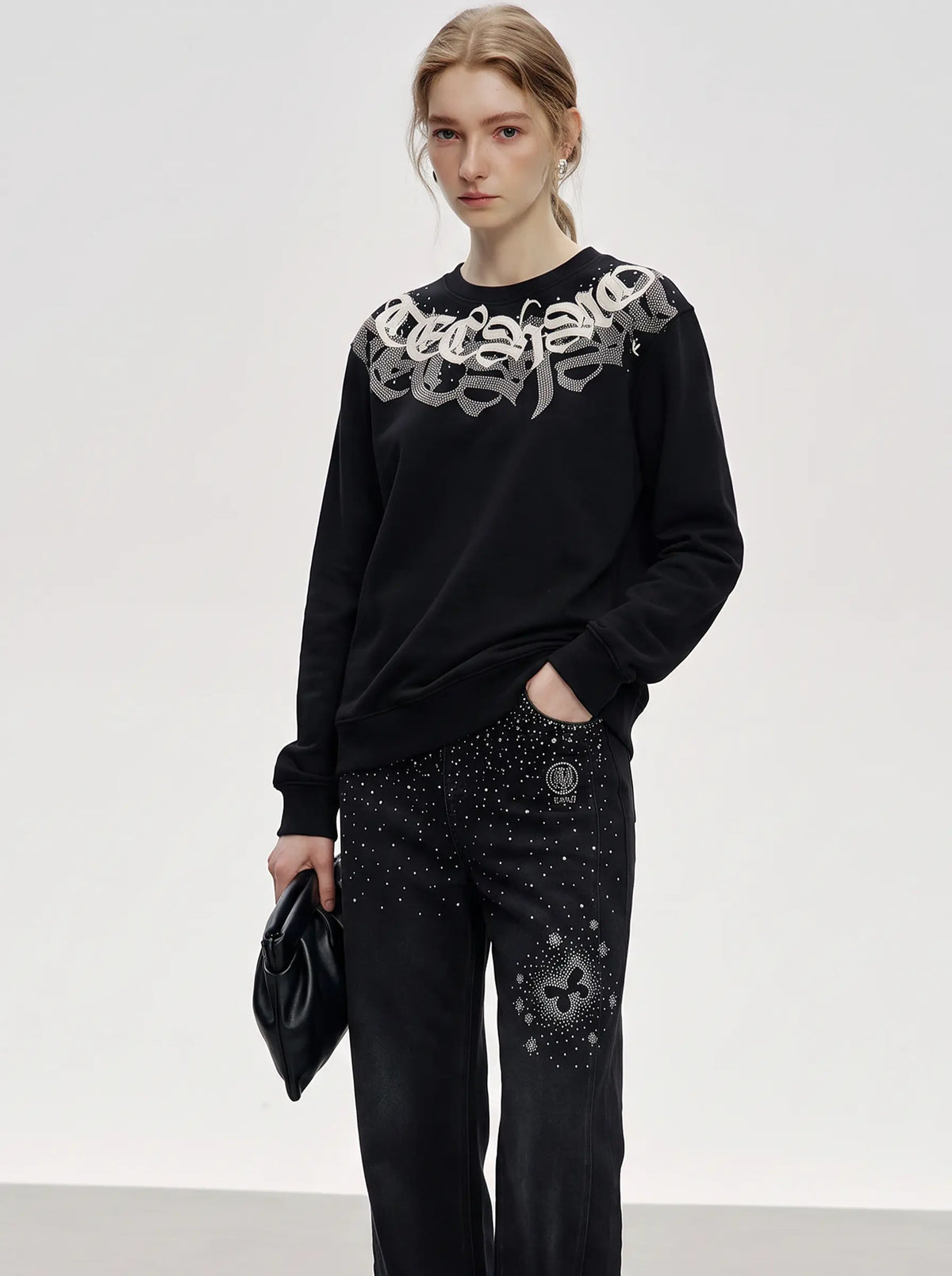 A model wearing a black long-sleeve sweatshirt with a large white graphic print across the chest, paired with black pants featuring silver rhinestone embellishments and a floral logo patch on the thigh, holding a black clutch bag against a light gray studio background. TCH Rhinestone Graphic Black Crewneck Sweatshirt