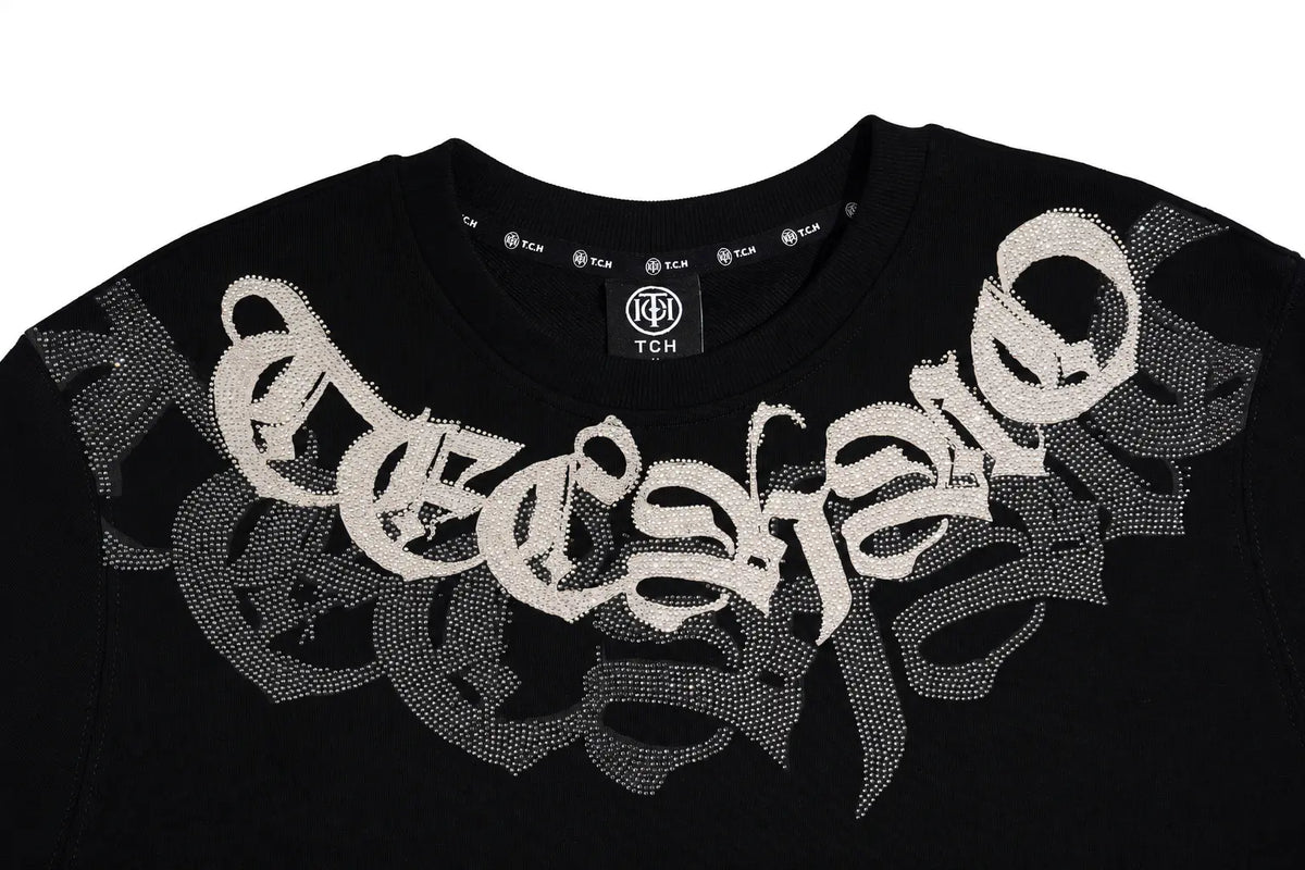 A close-up of a black TCH brand t-shirt featuring a large, textured white gothic-style 'eccentric' graphic across the chest, set against a plain white background. TCH Rhinestone Graphic Black Crewneck Sweatshirt