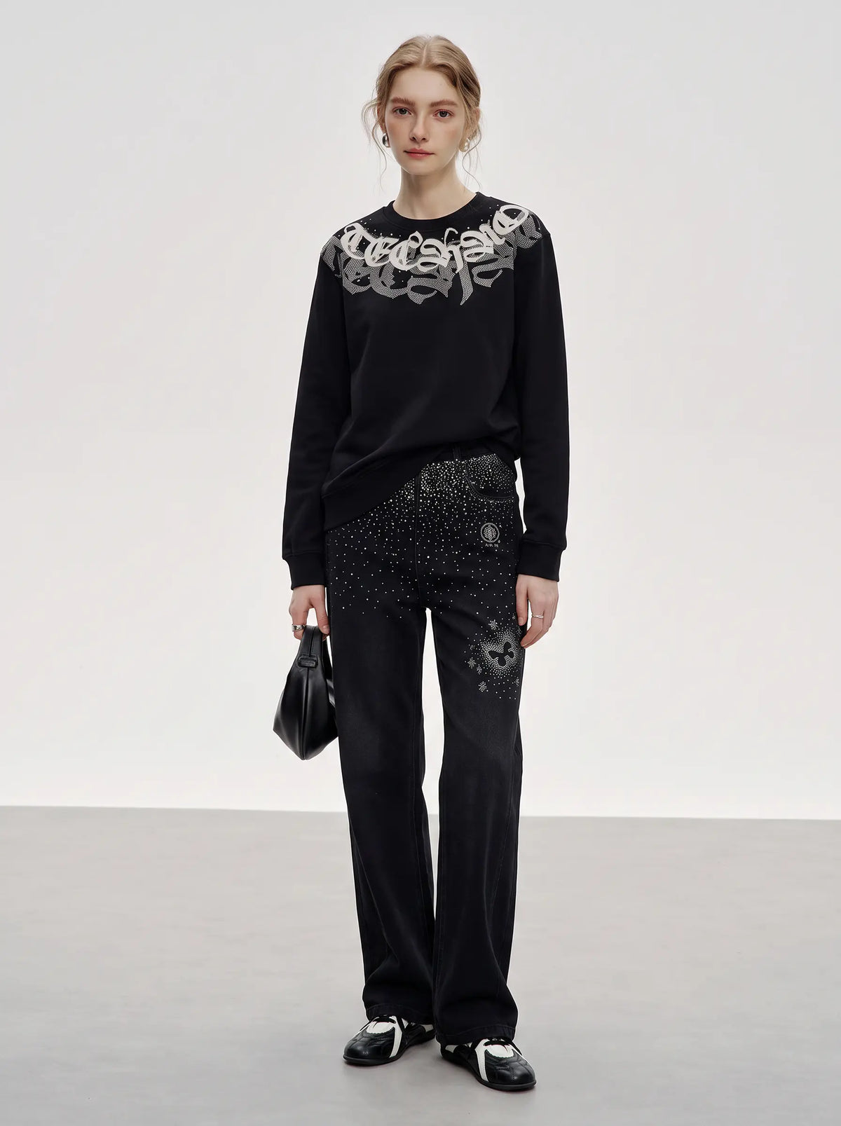 A model stands in a studio wearing a black long-sleeve top with a large silver graphic across the shoulders, black embellished pants, and black-and-white sneakers, while holding a small black handbag. TCH Rhinestone Graphic Black Crewneck Sweatshirt