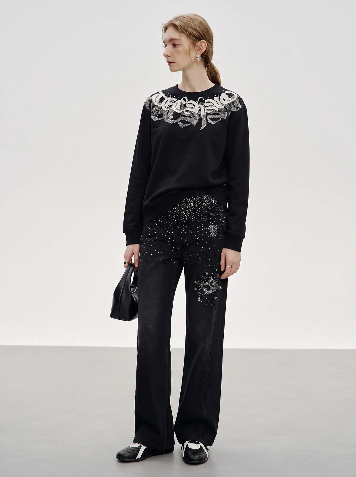 A model stands in a studio wearing a black long-sleeve sweatshirt with a large white graphic print across the chest, paired with black wide-leg pants featuring rhinestone embellishments and black-and-white sneakers. TCH Rhinestone Graphic Black Crewneck Sweatshirt