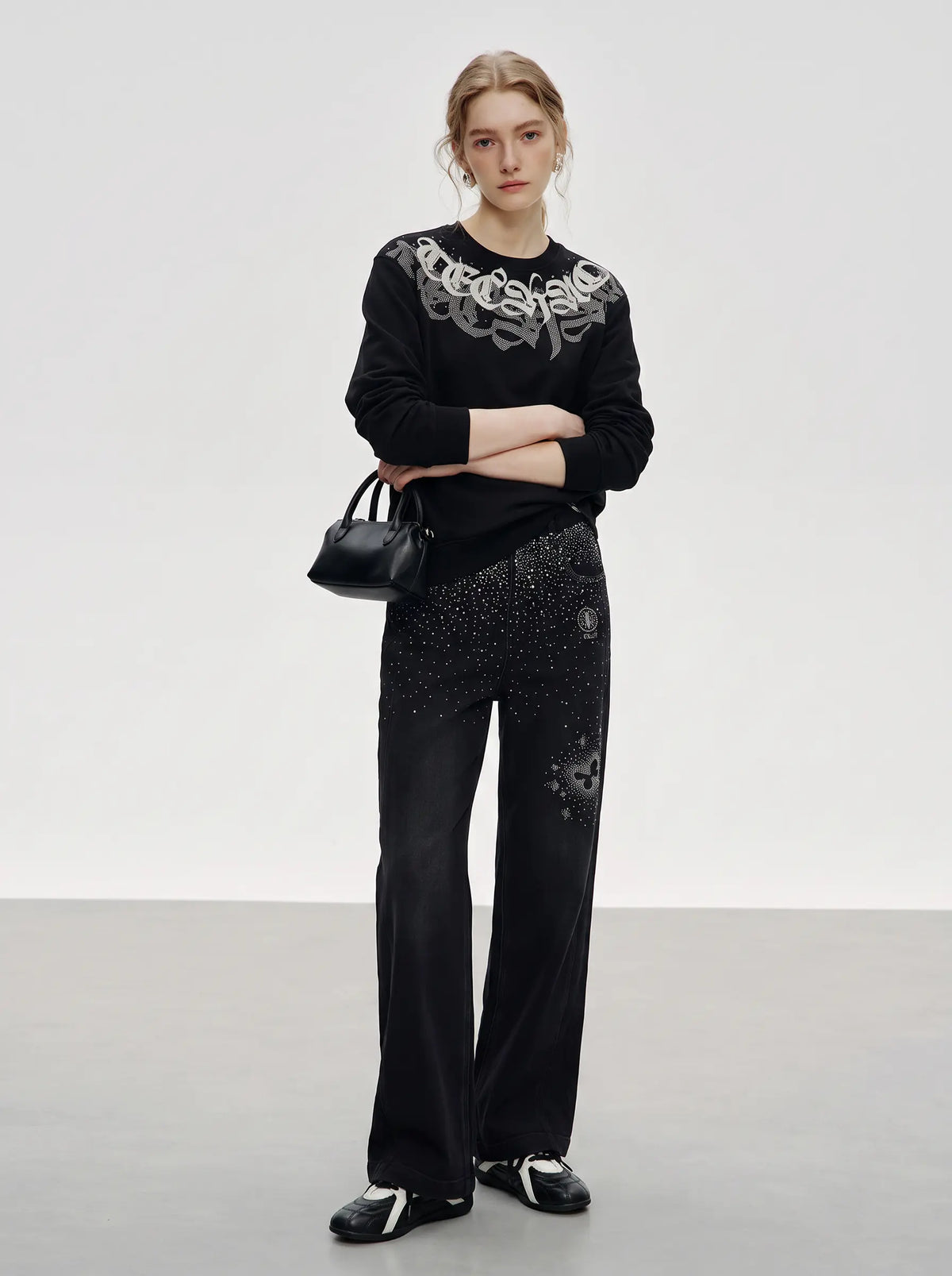 A model stands in a studio wearing a black long-sleeve top with a large silver graphic across the chest, black pants with sparkling embellishments, and black-and-white sneakers, while holding a small black handbag. TCH Rhinestone Graphic Black Crewneck Sweatshirt