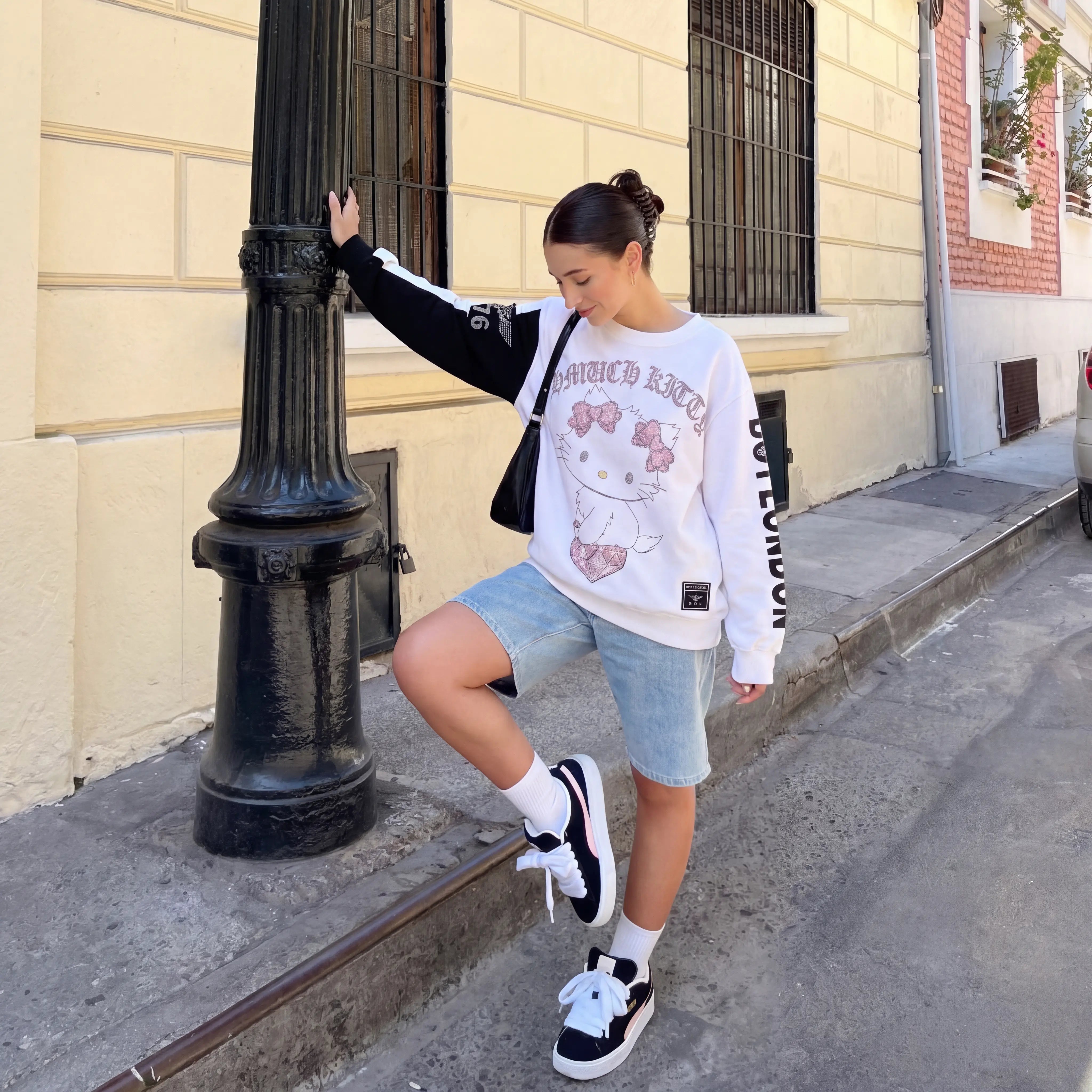 Woman in a white sweatshirt with cartoon character design, denim shorts, and black sneakers standing on a sidewalk. TCH Rhinestone Hello Kitty BOYLONDON Sweatshirt