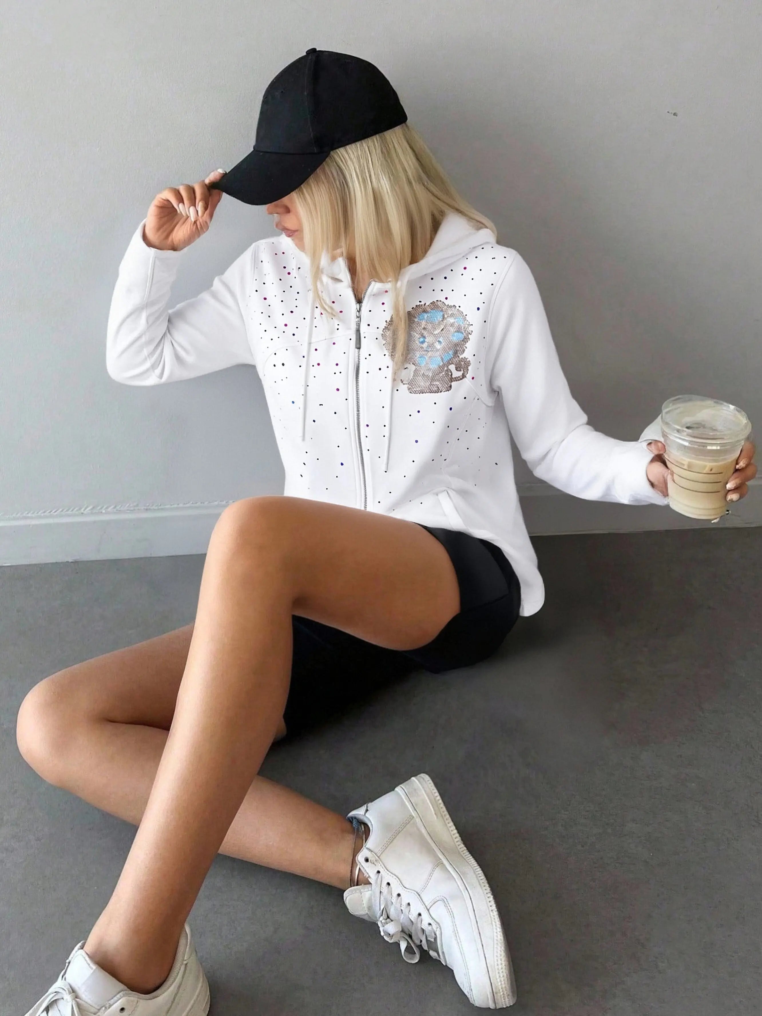 Person wearing a white hoodie with decorative elements, black shorts, and white sneakers, sitting on the floor holding a drink. TCH Rhinestone Lion & Horse Head Hoodie