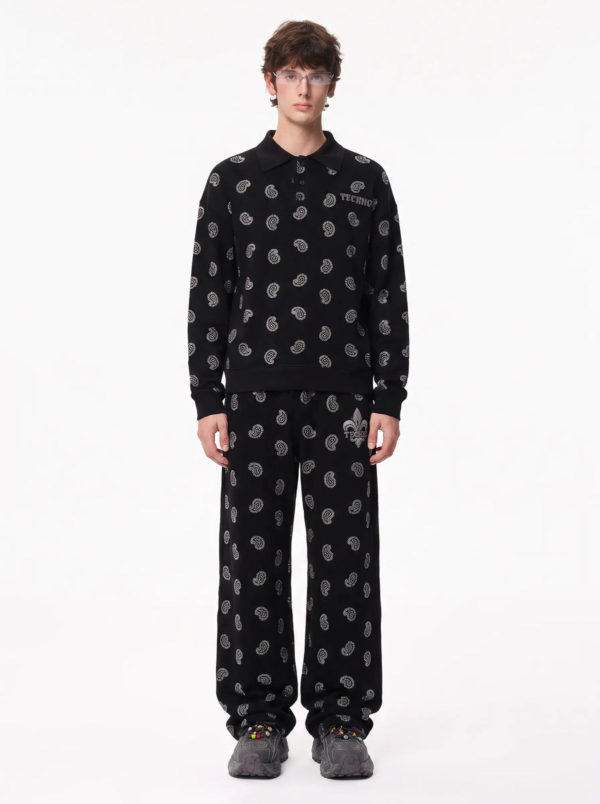 A model stands against a plain white studio background wearing a matching black two-piece set with a repeating gray paisley pattern. The set includes a long-sleeve collared top and wide-leg pants, with gray slippers on the feet. TCH Rhinestone Paisley Pattern Long Sleeve Polo Shirt