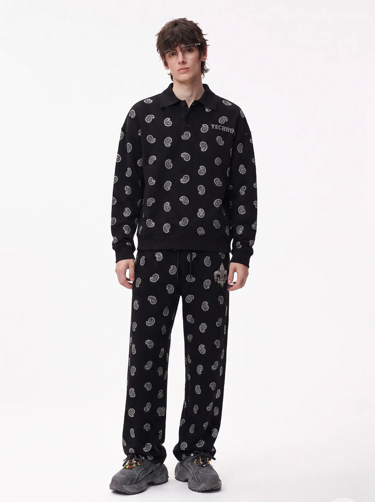 A model stands against a plain white studio background wearing a matching black two-piece tracksuit with a white abstract pattern, featuring a long-sleeve collared top and matching sweatpants. TCH Rhinestone Paisley Pattern Long Sleeve Polo Shirt