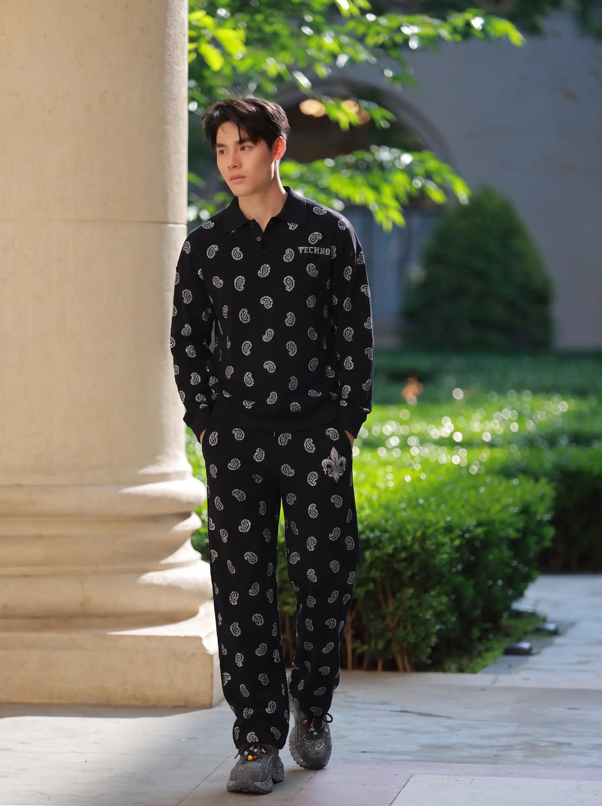 A man walking outdoors wearing a matching black tracksuit with a white paisley pattern, featuring a polo shirt and jogger pants, set against a background of green hedges and a stone column. TCH Rhinestone Paisley Pattern Long Sleeve Polo Shirt