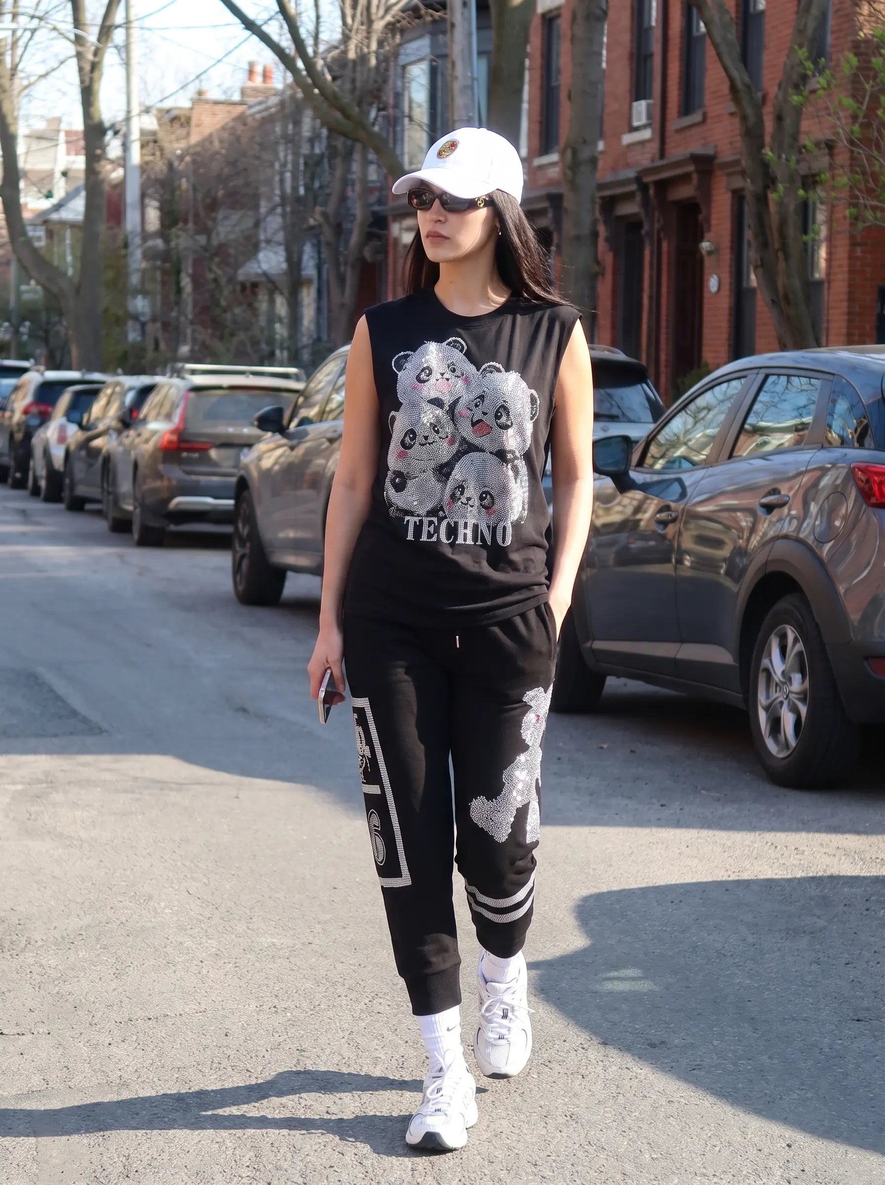 A woman walking down a city street wearing a black sleeveless Techno sweatshirt with a panda graphic, black jogger pants with white graphics, a white baseball cap, and white sneakers. TCH Rhinestone Panda Graphic Sleeveless T-Shirt