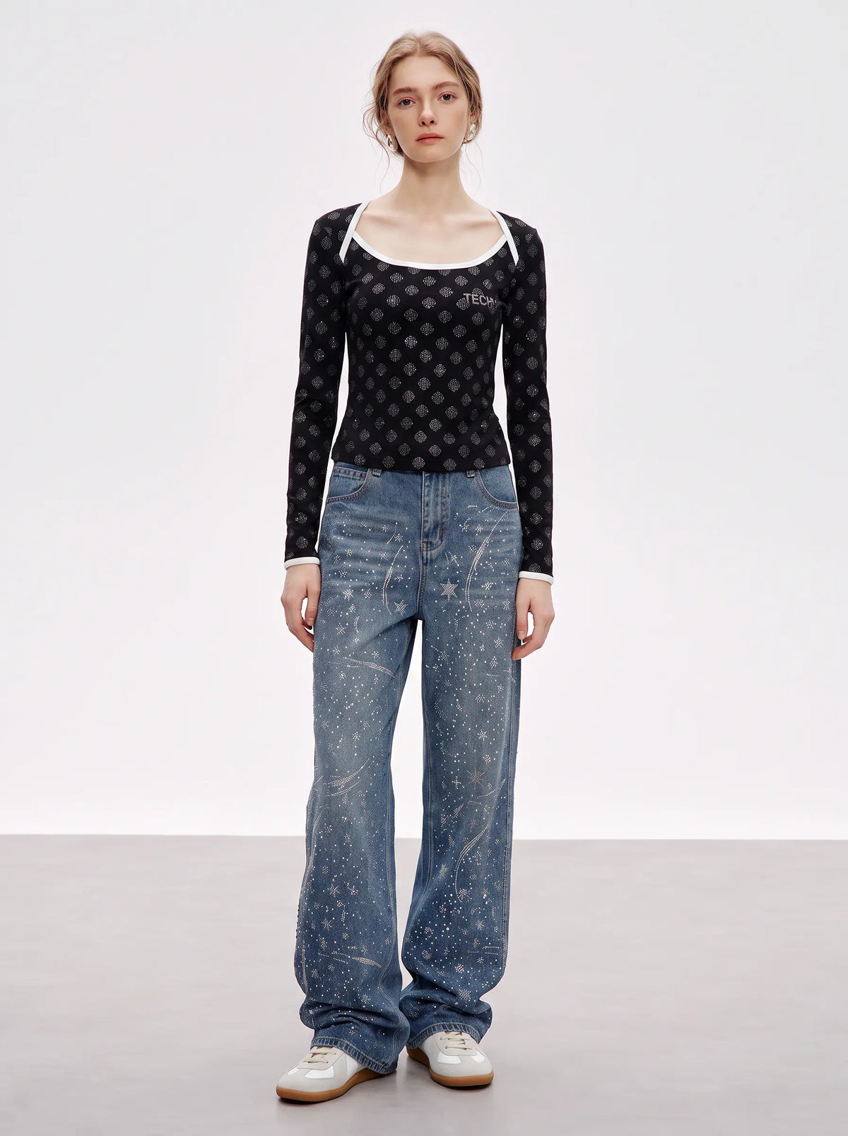A model stands against a light gray studio background wearing a black long-sleeve top with a small repeating white pattern, a white scoop-neck undershirt peeking out at the collar, wide-leg blue jeans with a white paint-splatter print, and white sneakers. TCH Rhinestone Pattern Long Sleeve Top