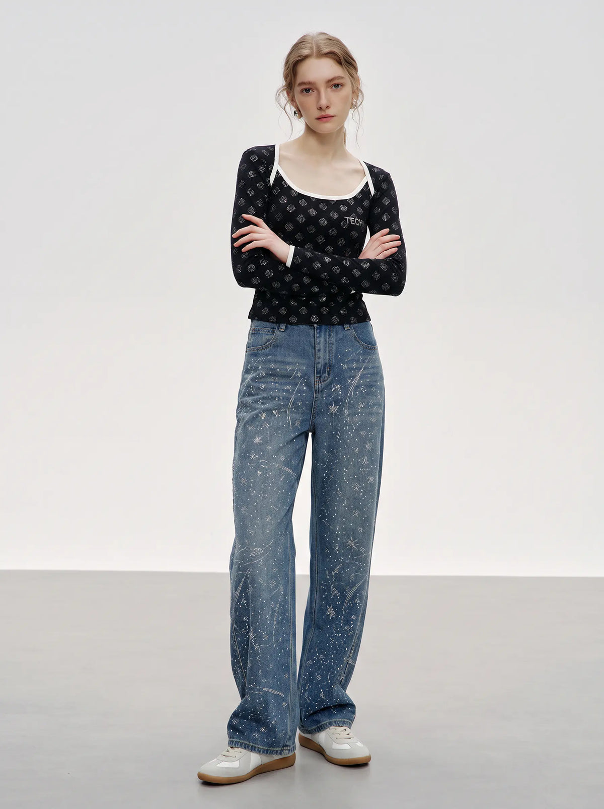 A model stands with arms crossed wearing a black long-sleeve top with a small white pattern, a white scoop-neck undershirt, wide-leg blue jeans with a white paint-splatter print, and white sneakers, set against a minimalist studio background. TCH Rhinestone Pattern Long Sleeve Top