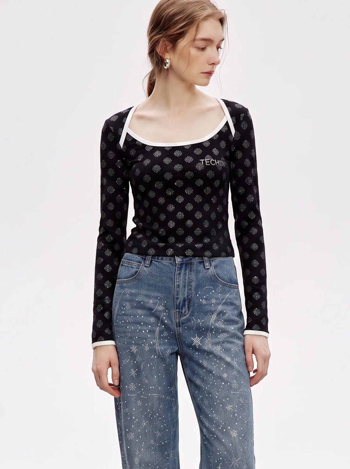 A model wearing a black long-sleeve scoop-neck top with a white geometric pattern and 'TECH' printed on the chest, paired with blue jeans, against a plain white studio background. TCH Rhinestone Pattern Long Sleeve Top