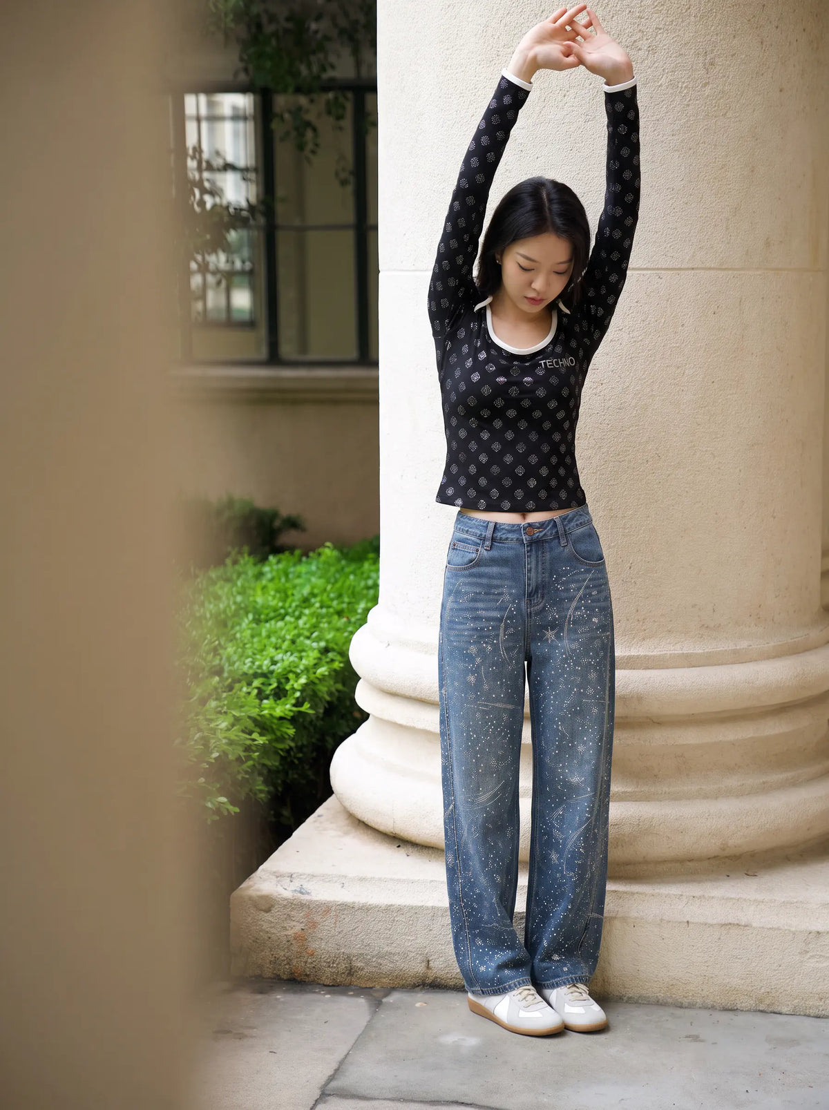 A woman stands outdoors in front of a large stone column, wearing a fitted black long-sleeve top with a small repeating light pattern, loose blue jeans, and white sneakers. TCH Rhinestone Pattern Long Sleeve Top