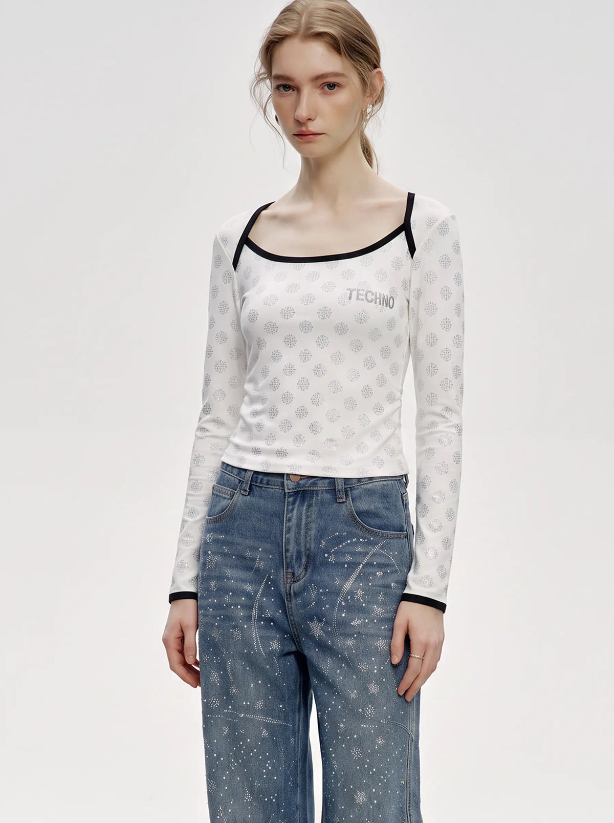 A model wearing a white long-sleeve crop top with a subtle geometric pattern and black trim, paired with blue jeans, against a plain light gray studio background. TCH Rhinestone Pattern Long Sleeve Top