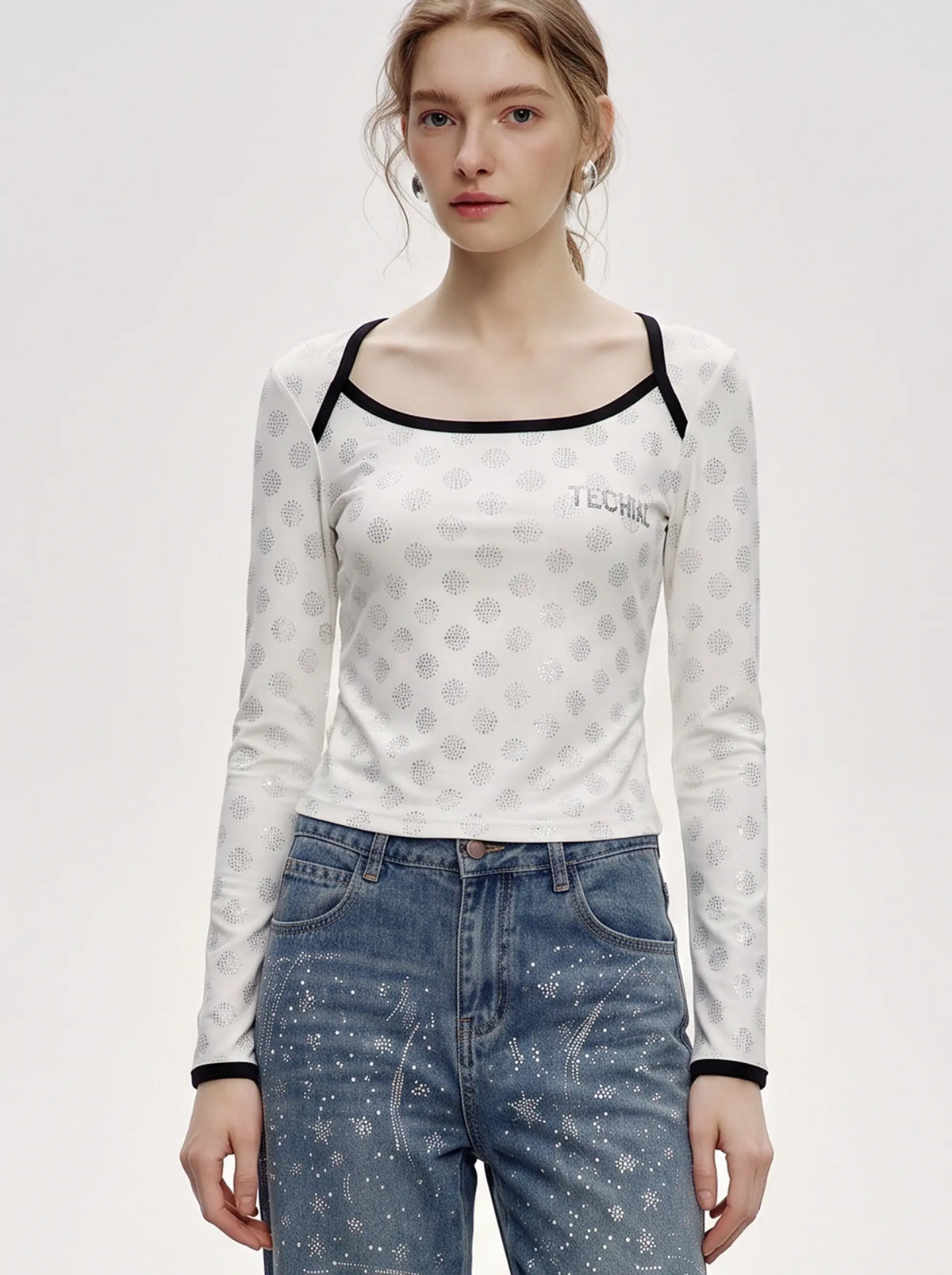A woman modeling a white long-sleeve crop top with a gray circular pattern, black trim, and a small 'TECHNIC' logo on the chest, paired with blue jeans against a light gray studio background. TCH Rhinestone Pattern Long Sleeve Top