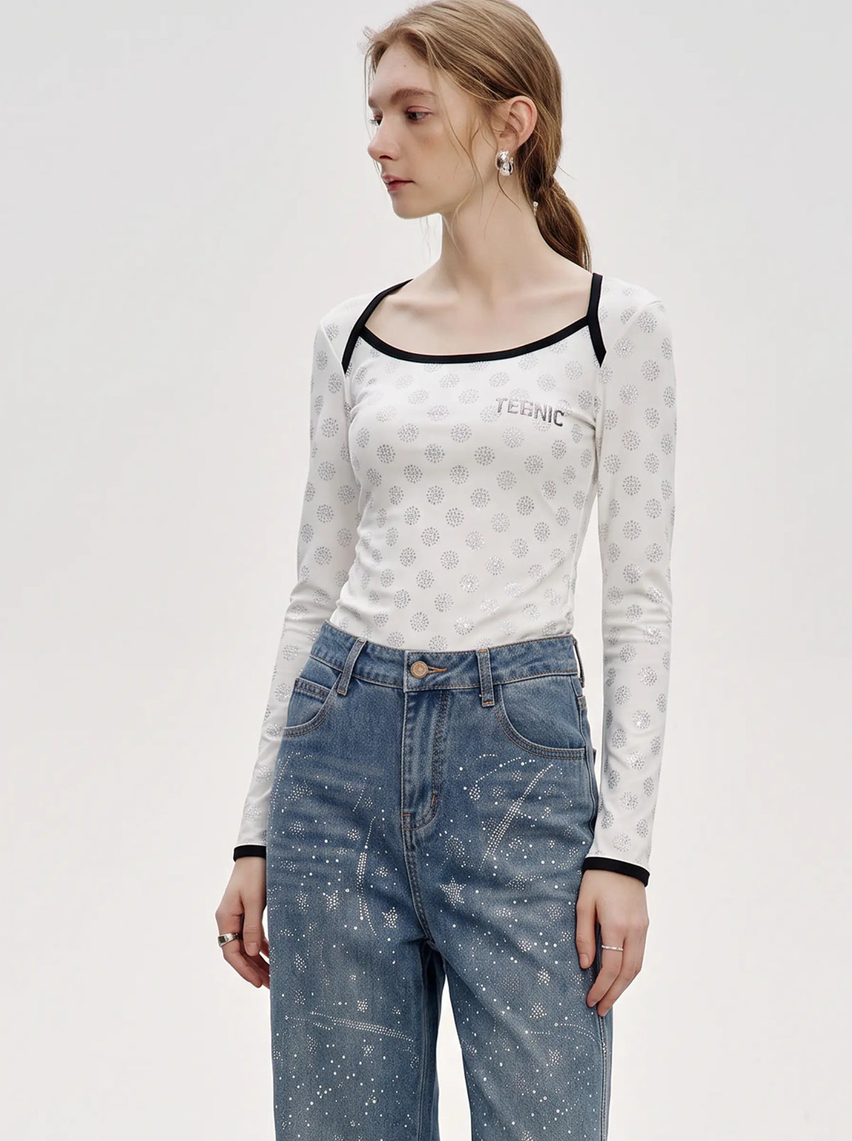 A woman modeling a white long-sleeve top with a subtle gray geometric pattern, black trim along the scoop neckline and cuffs, and 'TEENIC' printed on the chest, paired with blue jeans against a light gray studio background. TCH Rhinestone Pattern Long Sleeve Top