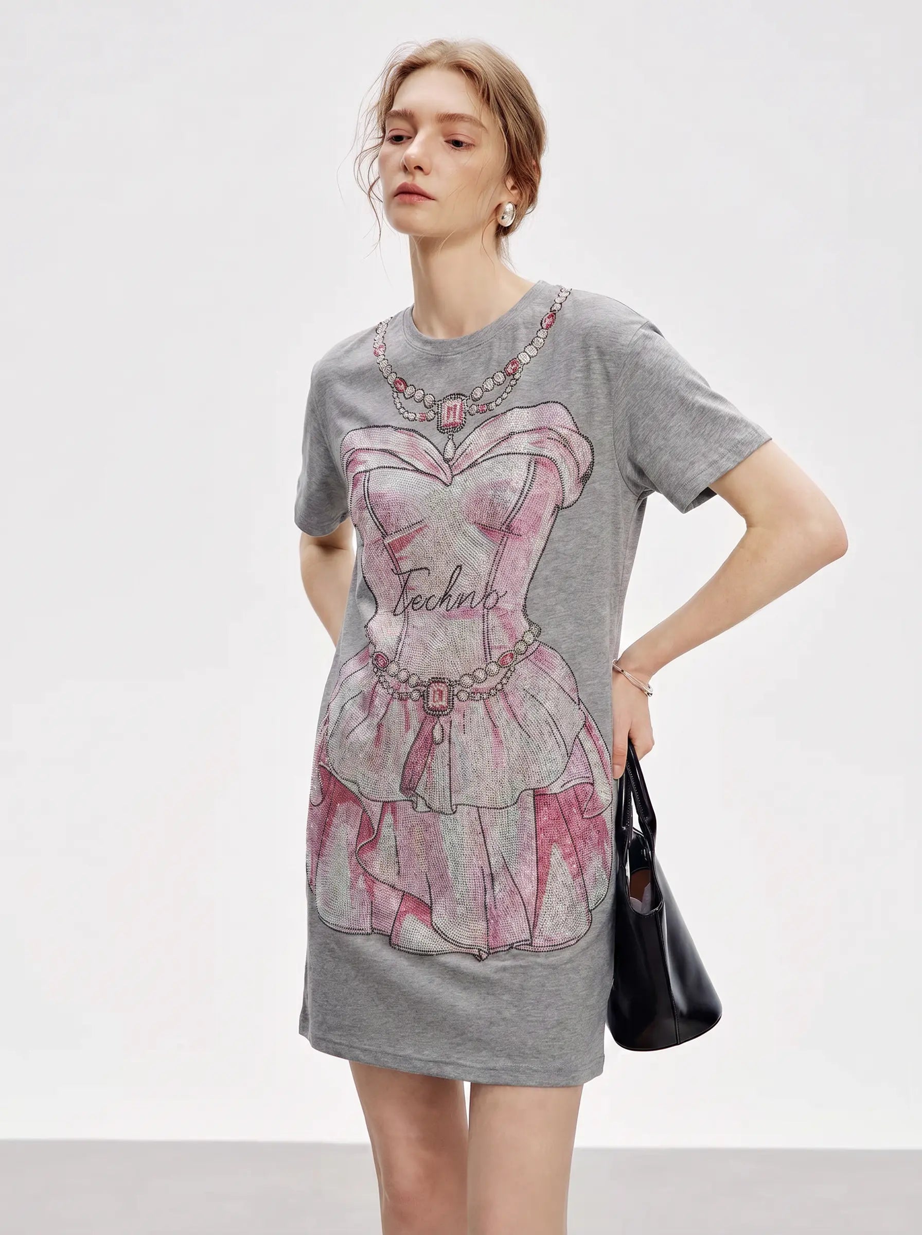 A woman modeling a heather gray short-sleeve T-shirt dress featuring a large pink and gray graphic of a corset dress with the word 'Teacher' printed on the front, holding a black handbag against a light gray studio background. TCH Rhinestone Pearl Corset Graphic Mini Dress
