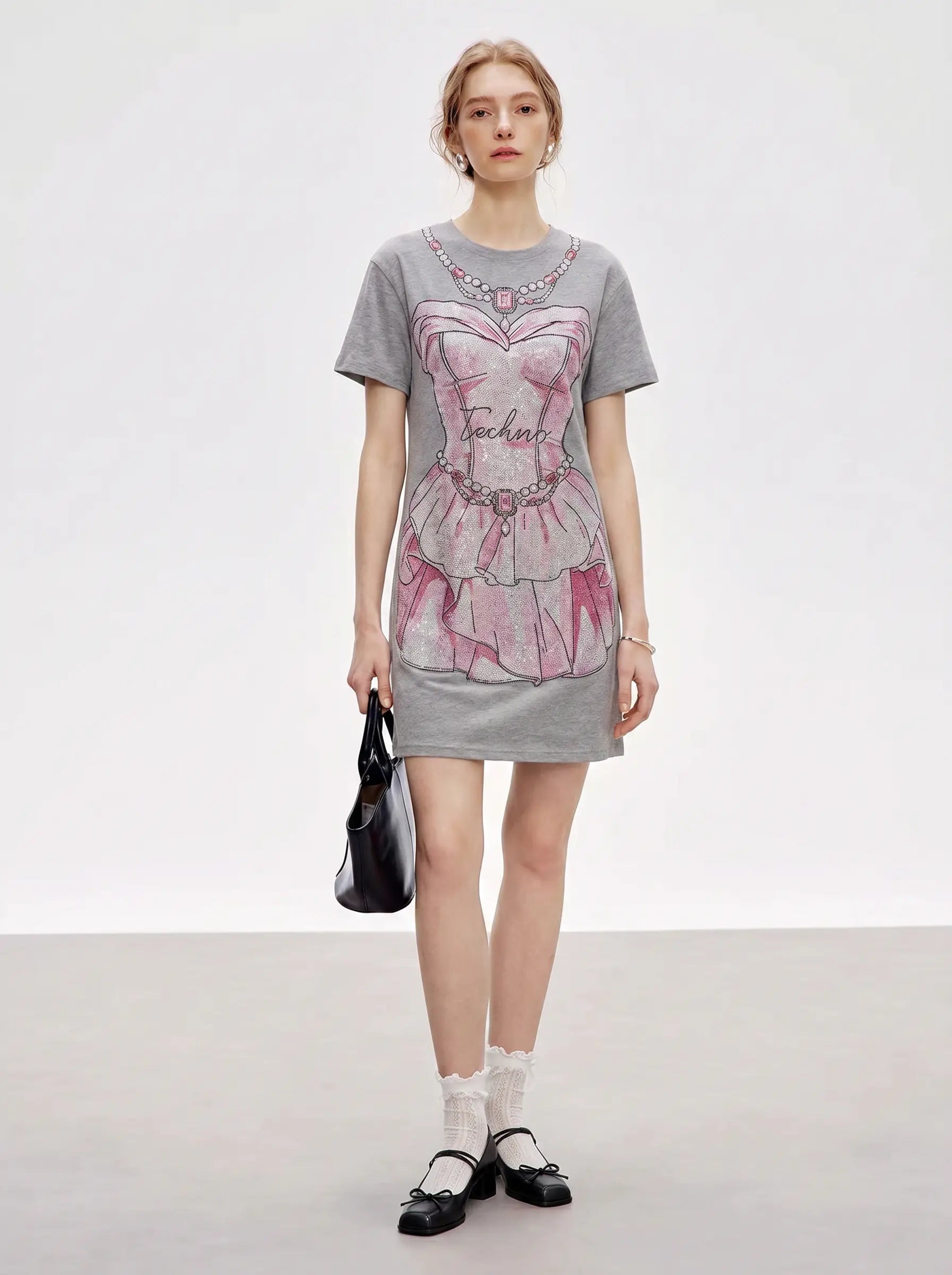 A model stands against a light gray studio background wearing a gray short-sleeve T-shirt dress with a pink and white corset-style graphic print. She is holding a black handbag and wearing white socks and black Mary Jane shoes. TCH Rhinestone Pearl Corset Graphic Mini Dress