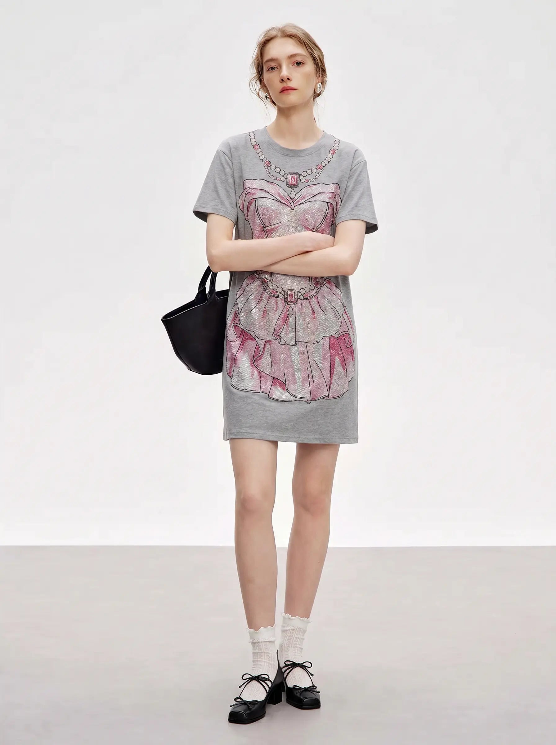 A model stands in a studio wearing a gray oversized graphic T-shirt dress with a pink and white ruffled dress illustration, paired with white socks, black Mary Jane shoes, and a black handbag. TCH Rhinestone Pearl Corset Graphic Mini Dress