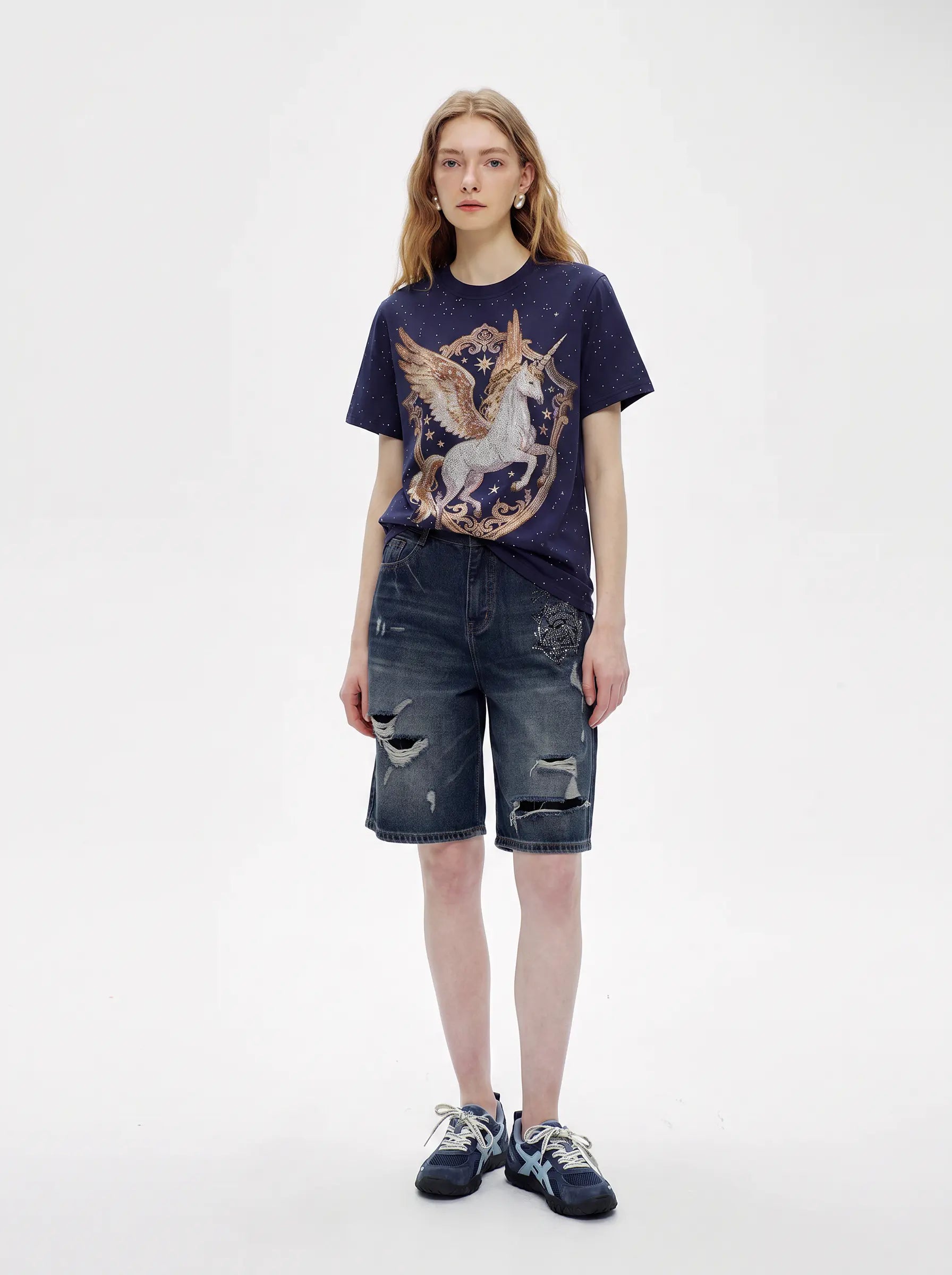 A model stands against a light gray studio background wearing a navy graphic T-shirt with a winged unicorn print, distressed dark denim shorts, and dark sneakers. TCH Rhinestone Pegasus Wing Graphic Navy Blue T-Shirt