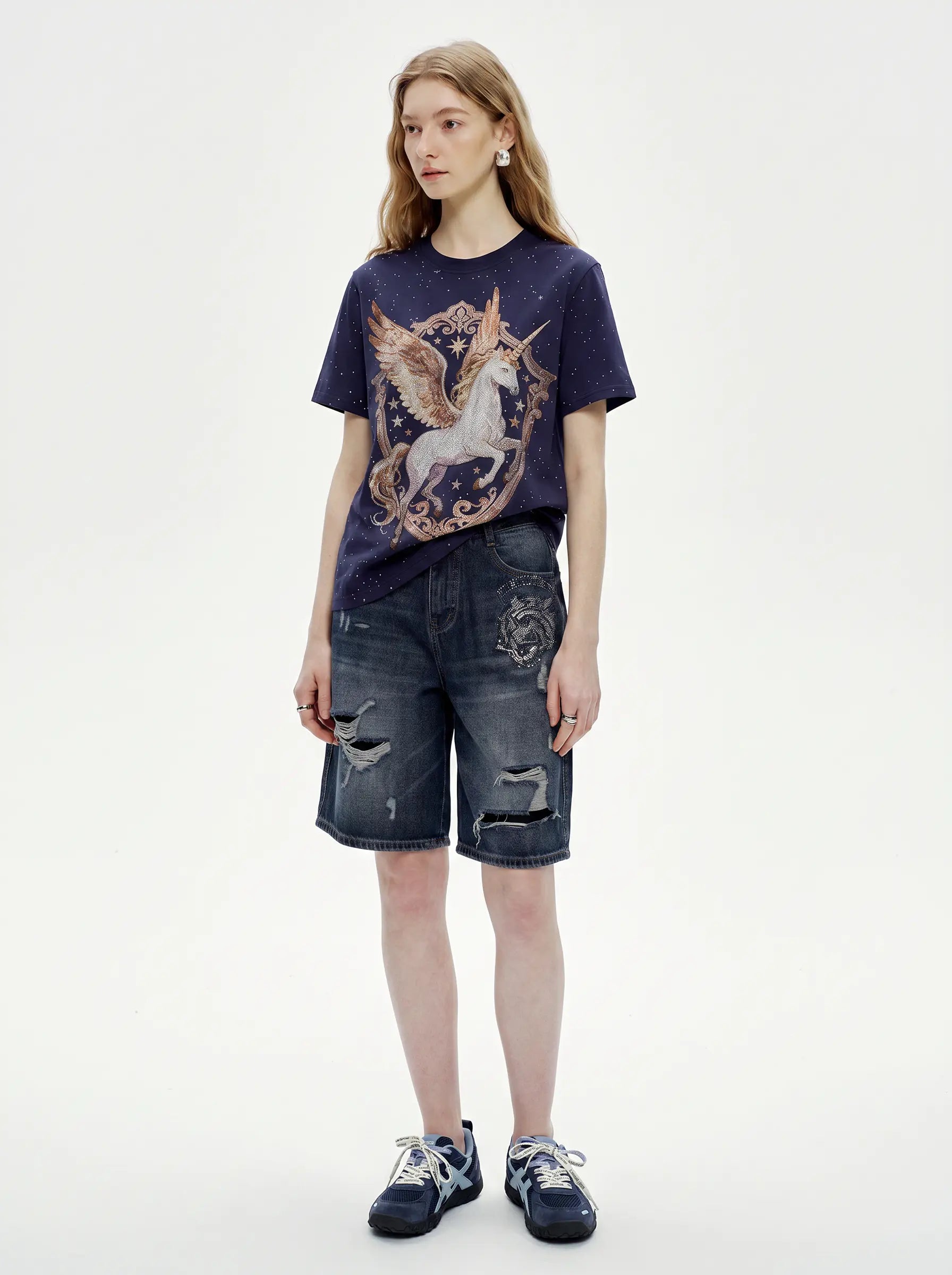 A model stands against a plain light background wearing a dark navy graphic T-shirt with a unicorn print, distressed denim shorts, and dark sneakers. TCH Rhinestone Pegasus Wing Graphic Navy Blue T-Shirt