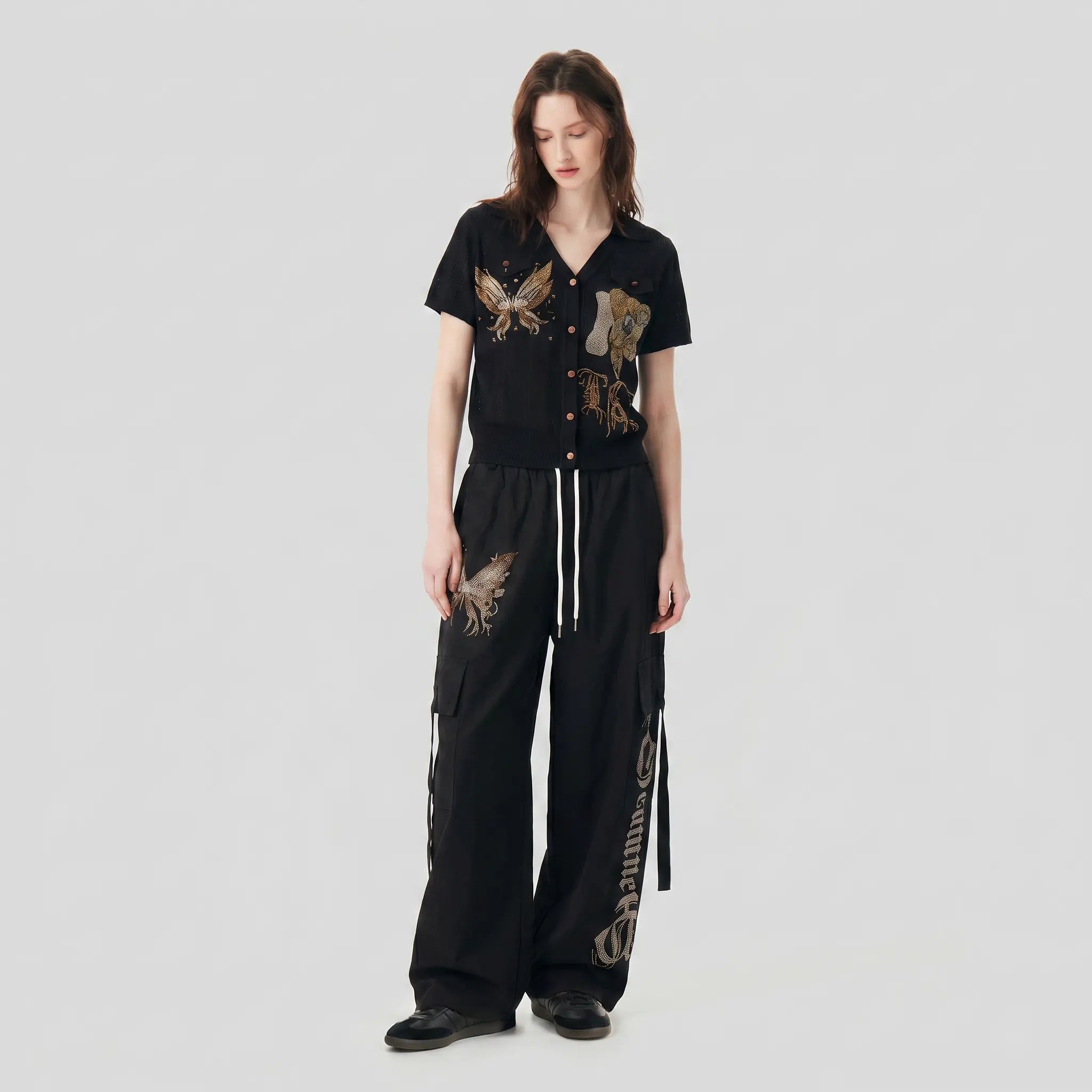 A model stands against a light gray studio background wearing a black short-sleeve button-front top with embroidered eagle motifs and matching black wide-leg pants with white drawstrings and side lettering. TCH Rhinestone Phoenix Cargo Wide-Leg Pants