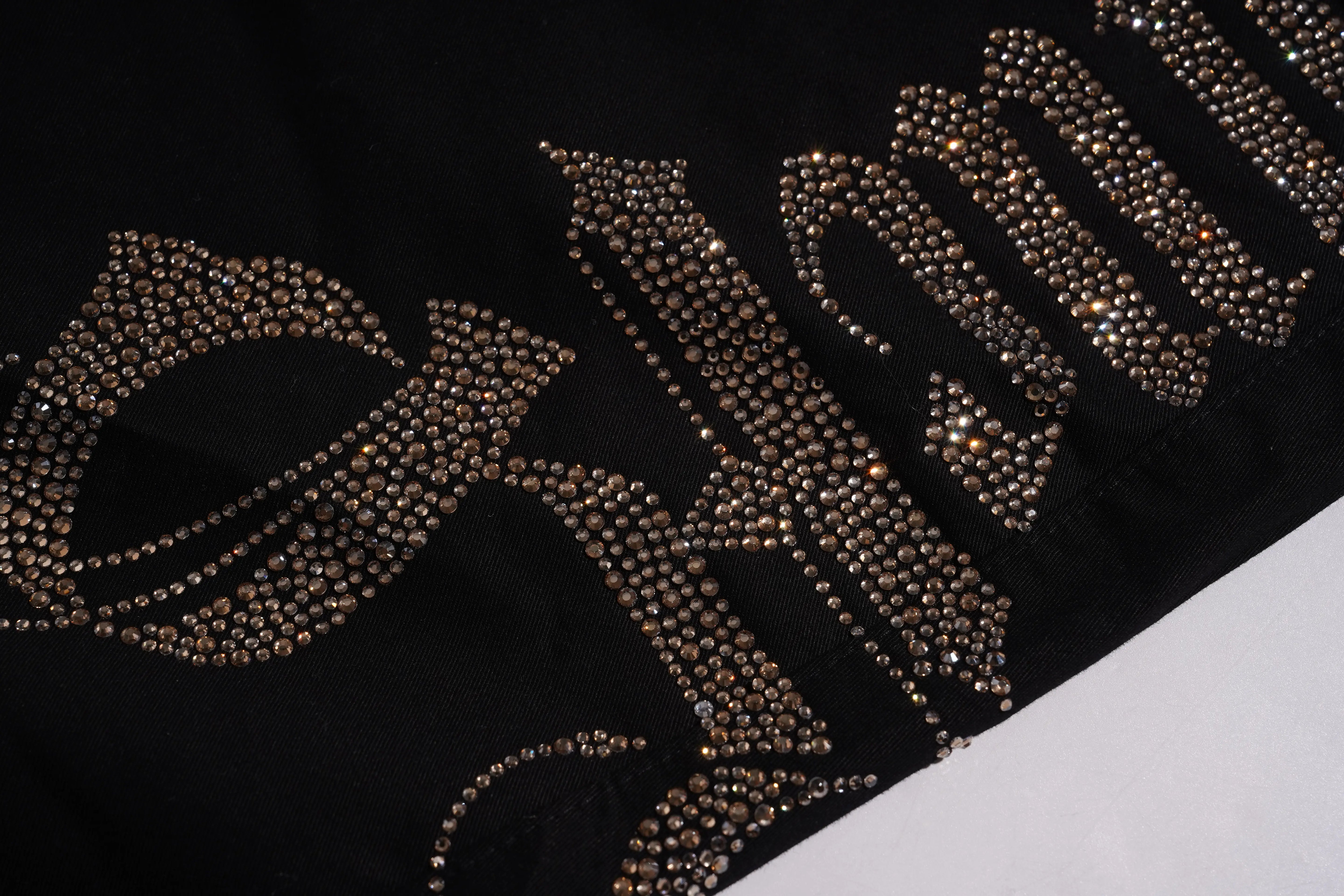 A close-up of a black garment featuring large, ornate lettering embellished with rows of metallic gold and silver rhinestones. TCH Rhinestone Phoenix Cargo Wide-Leg Pants