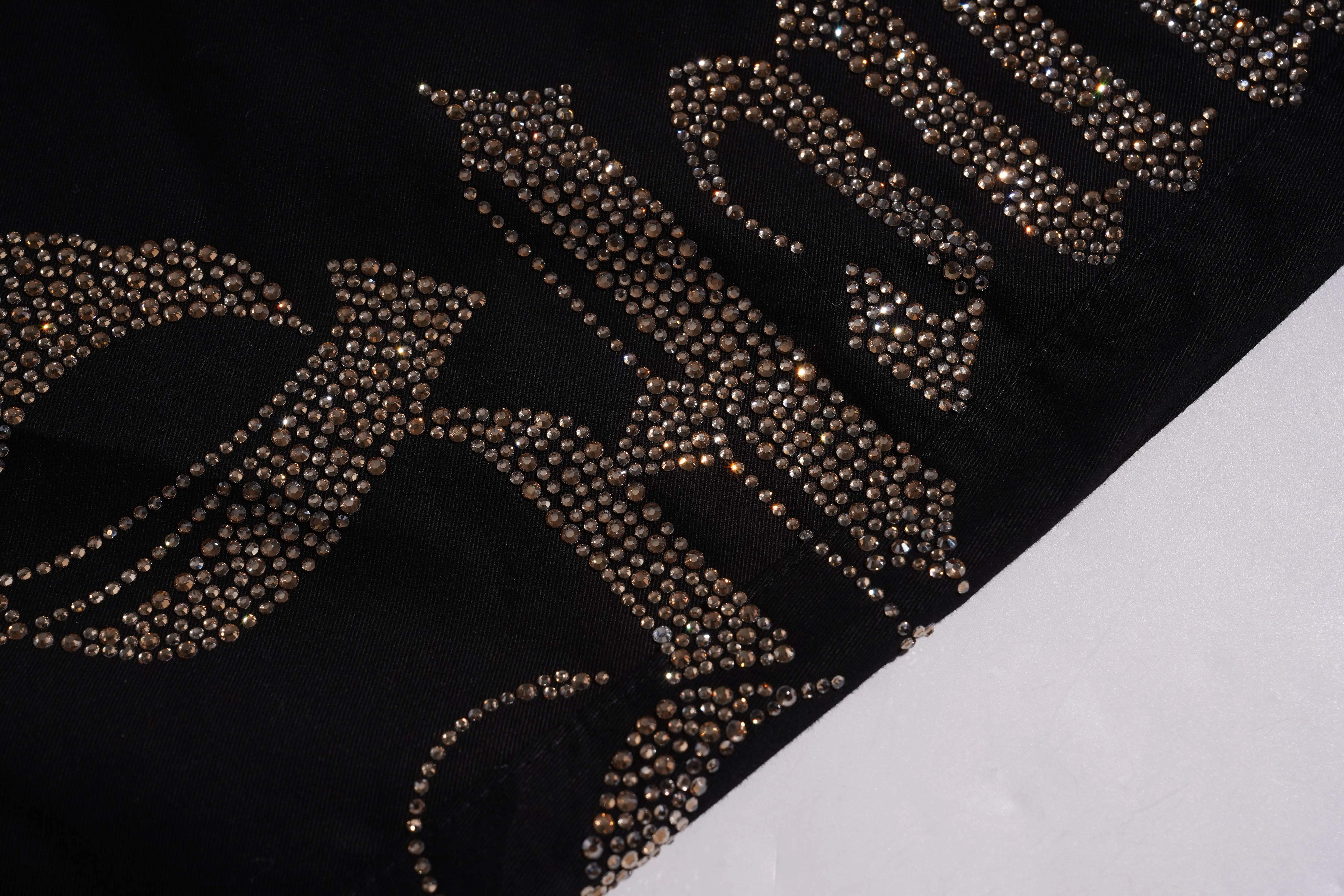 A close-up of a black fabric surface decorated with large, ornate lettering made of metallic gold and silver rhinestones, with a plain white surface visible in the lower right corner. TCH Rhinestone Phoenix Cargo Wide-Leg Pants