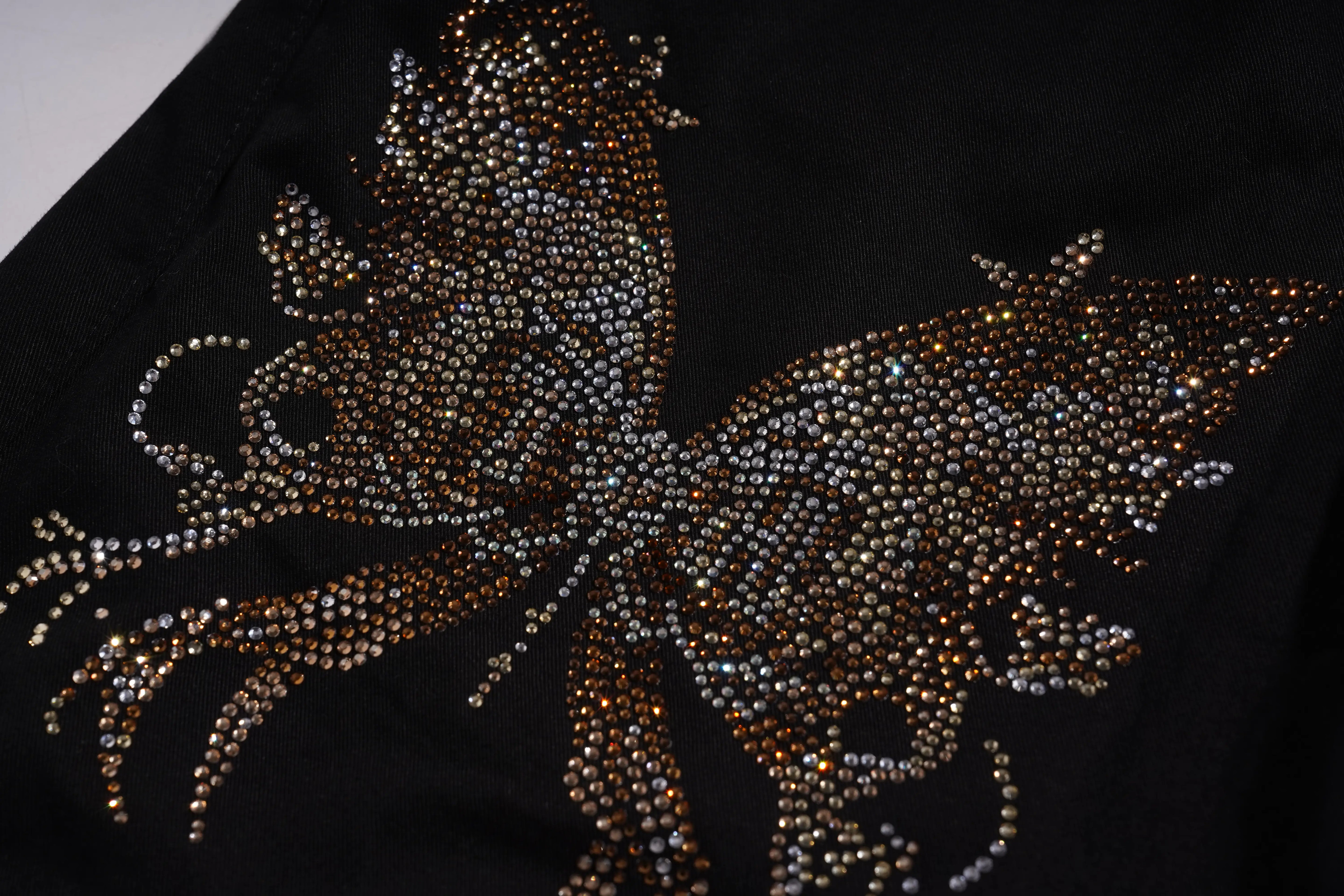 A close-up of a black garment featuring a large, intricate butterfly embellished with shimmering rhinestones in gold, silver, and bronze tones. TCH Rhinestone Phoenix Cargo Wide-Leg Pants