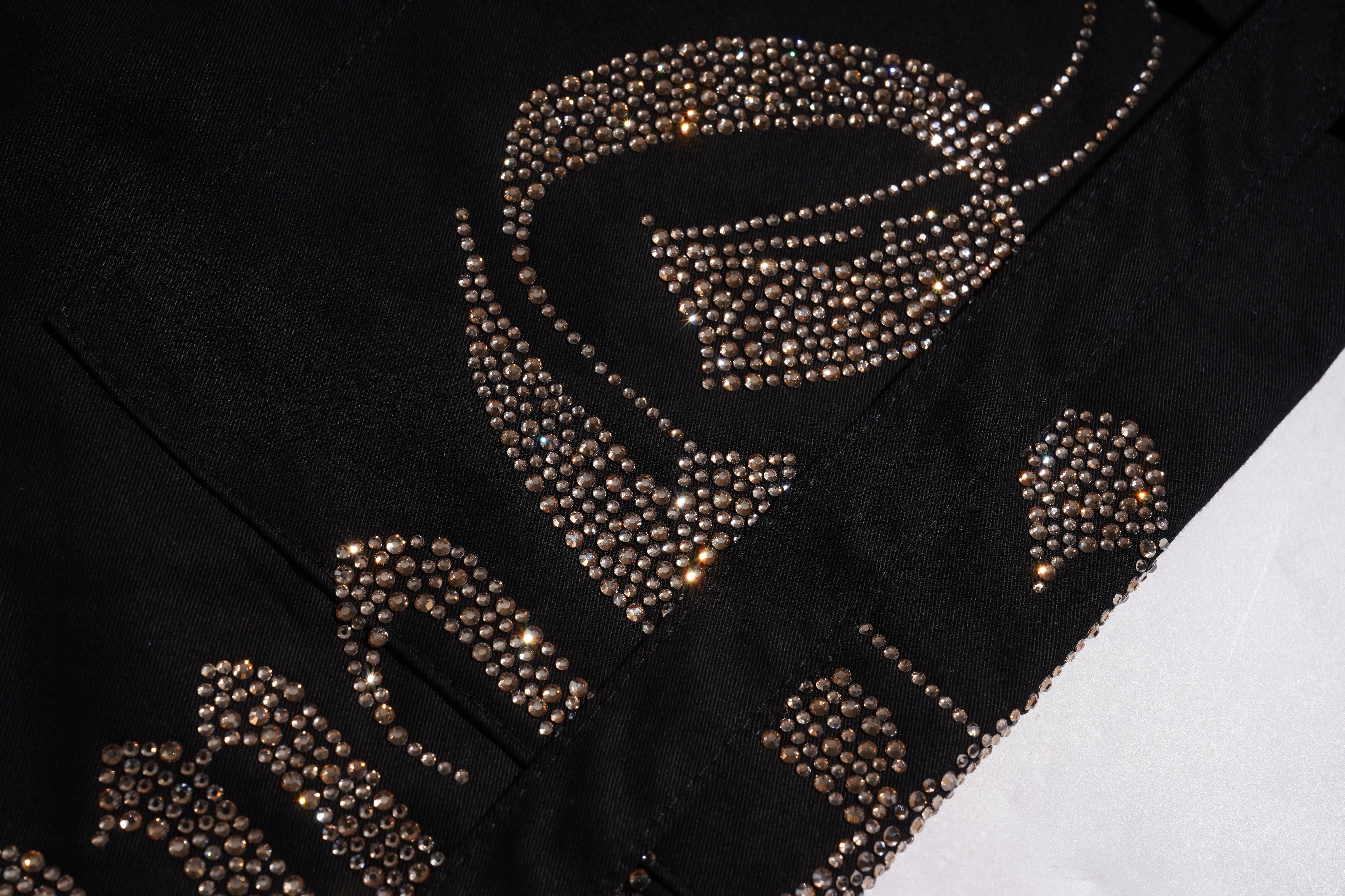 Close-up of a black garment featuring large metallic gold rhinestone embellishments in an abstract pattern, with a white surface visible in the lower right corner. TCH Rhinestone Phoenix Cargo Wide-Leg Pants