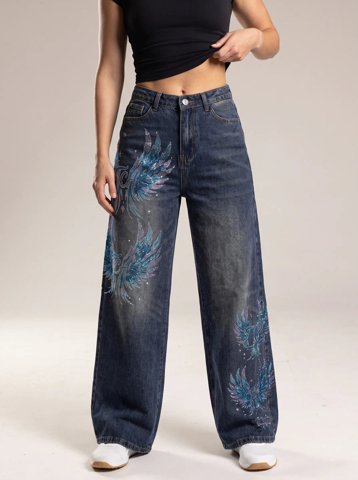 A model wearing a pair of wide-leg dark blue denim jeans with blue and purple feather patterns printed on the front, paired with a black crop top and white sneakers. TCH Rhinestone Phoenix Wide-Leg Jeans