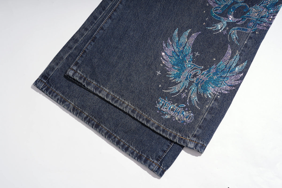 Close-up of folded dark blue denim jeans on a white background, featuring a large iridescent blue and purple winged graphic with small star details and stylized text near the hem. TCH Rhinestone Phoenix Wide-Leg Jeans