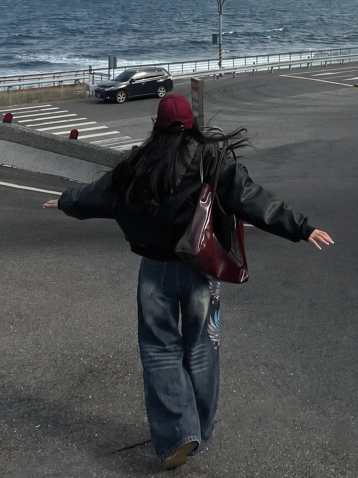 A person seen from behind walking on a coastal road with the ocean in the background, wearing a black leather jacket, wide-leg blue jeans, a burgundy beanie, and carrying a large burgundy shoulder bag. TCH Rhinestone Phoenix Wide-Leg Jeans