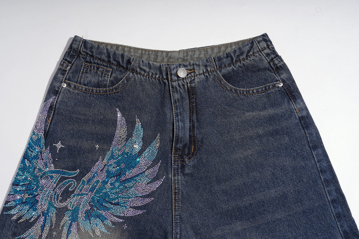 A close-up of a dark blue denim skirt featuring a large, glittery rhinestone wing graphic in shades of blue, purple, and silver on the left leg, set against a plain white background. TCH Rhinestone Phoenix Wide-Leg Jeans