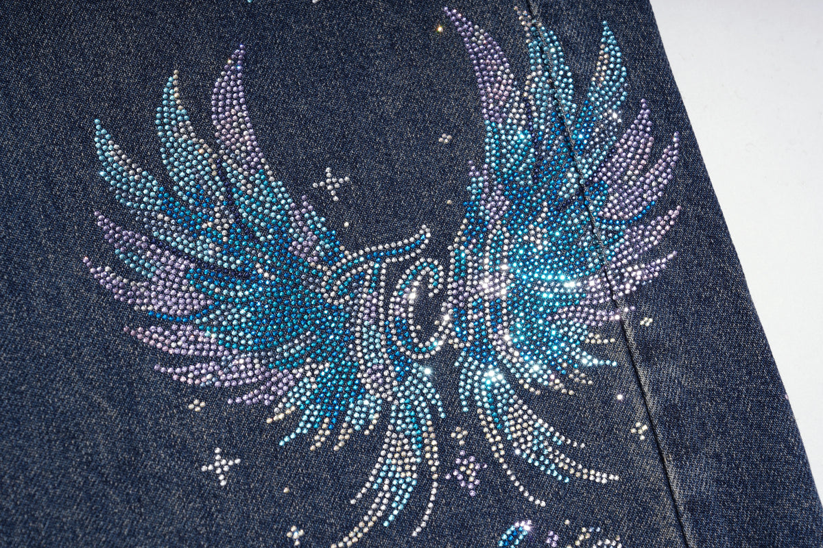 Close-up of a dark blue denim garment featuring a large rhinestone wing graphic in blue, purple, and silver, with small star accents. TCH Rhinestone Phoenix Wide-Leg Jeans