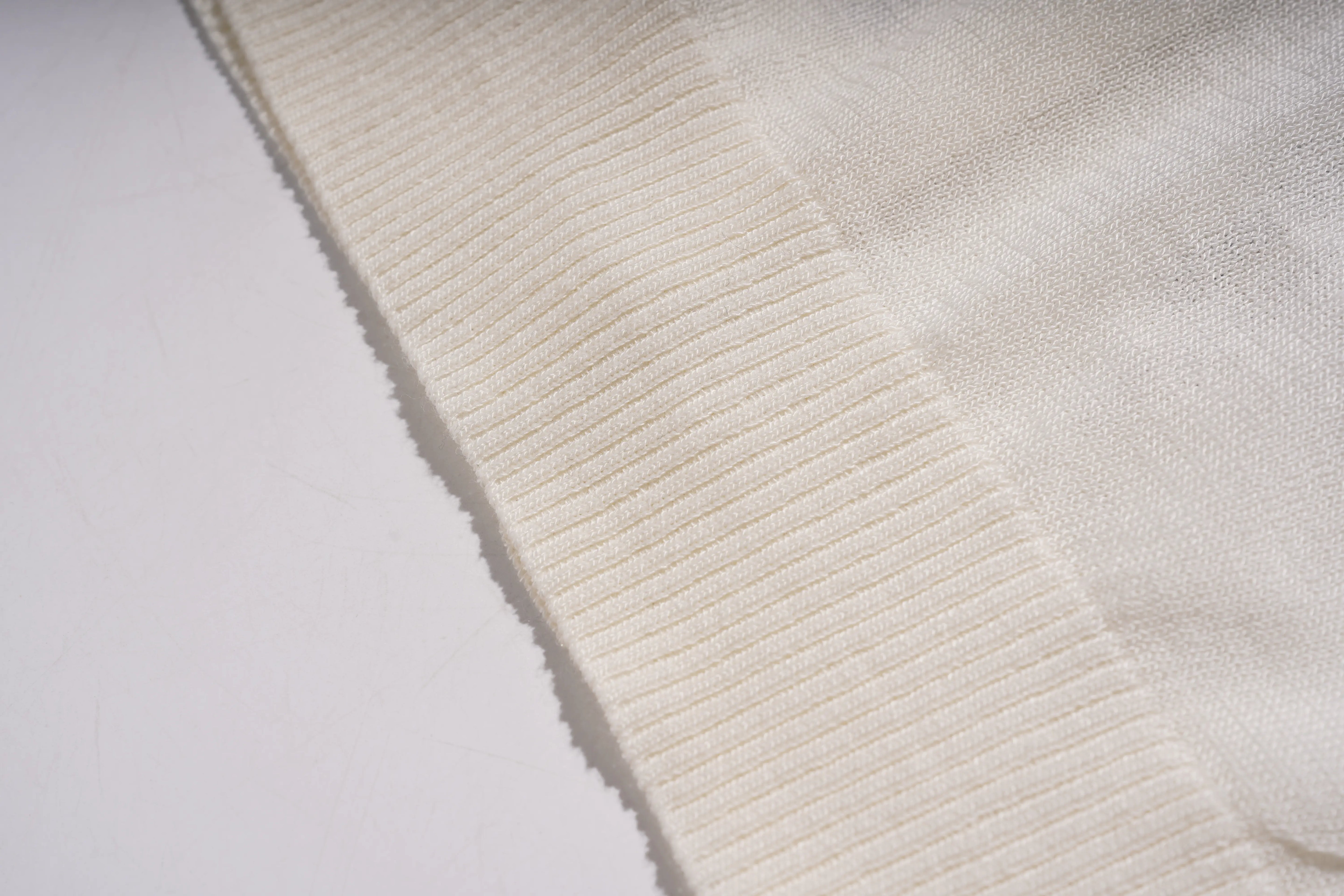 A close-up of a cream-colored ribbed knit fabric, showing the detailed texture of the ribbed edge and the smooth knit body, laid diagonally on a white surface. TCH Rhinestone Phoenix Wings Button Knit Top